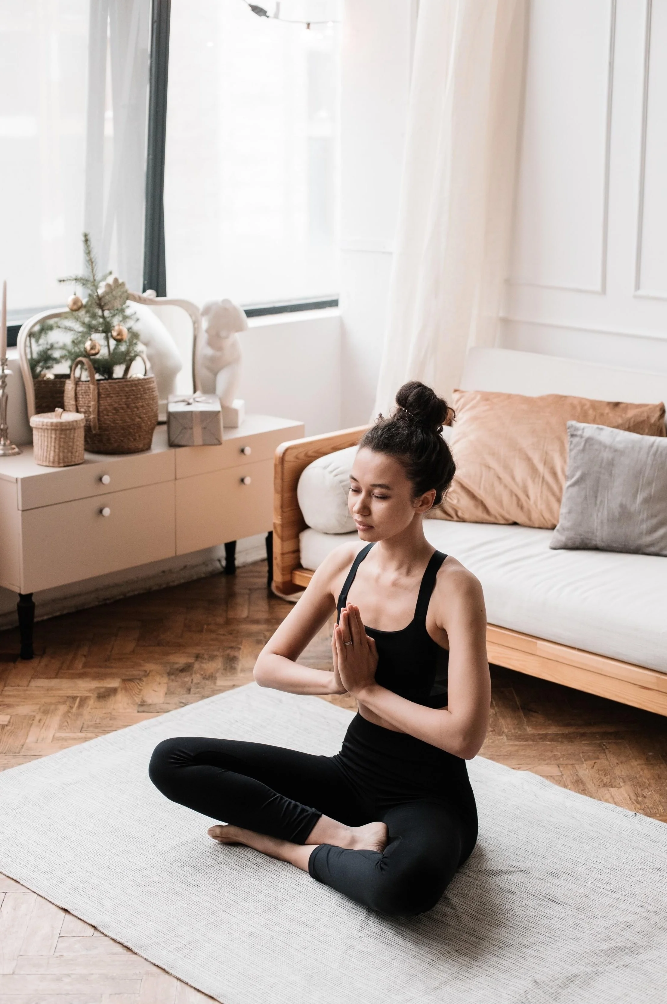 A woman practicing yoga indoors on a white mat, sitting cross-legged with hands in a prayer position, eyes closed, and a calm expression.