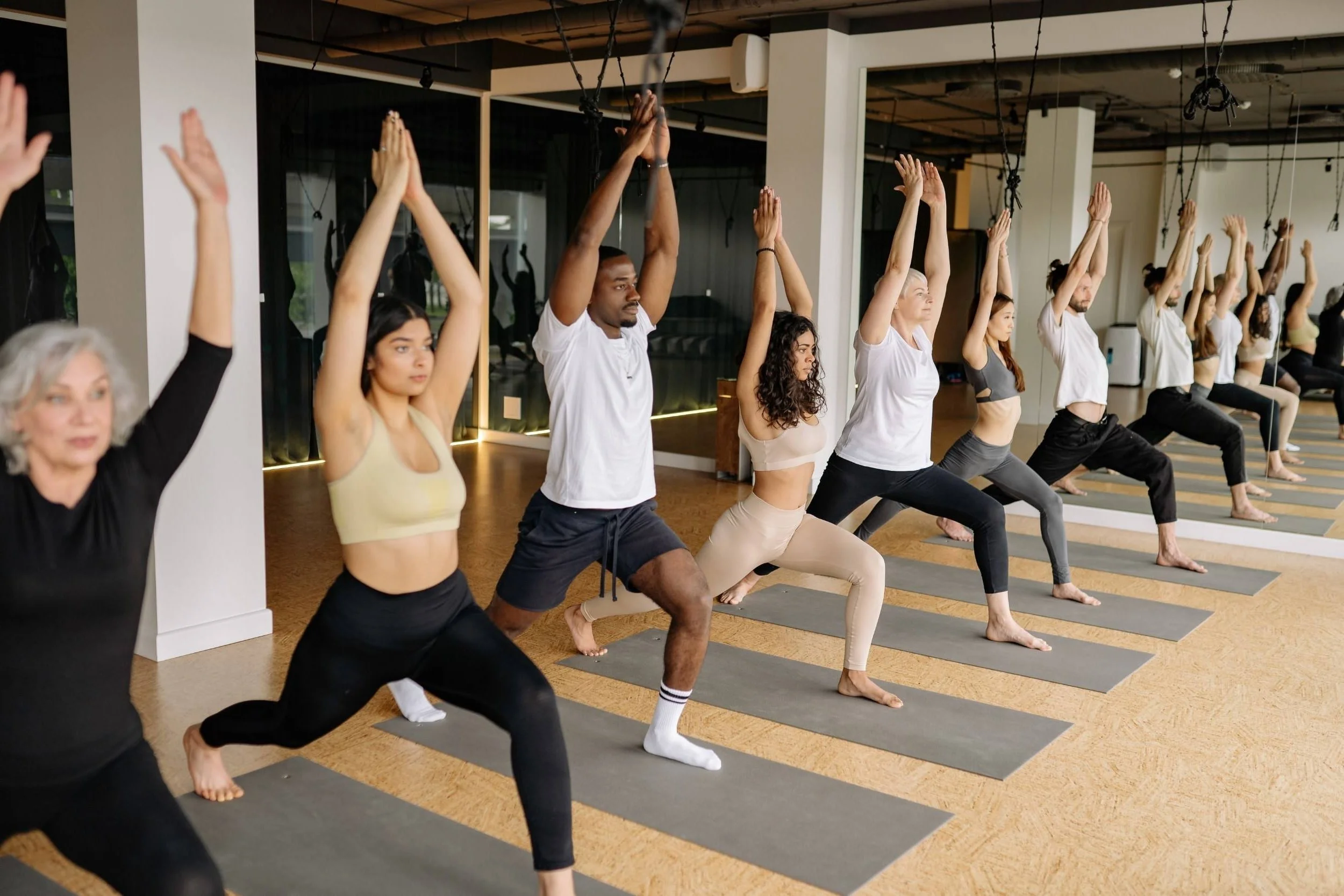 A diverse group of people practicing yoga in a studio, standing in a Warrior pose with their arms raised overhead and feet wide apart on yoga mats.