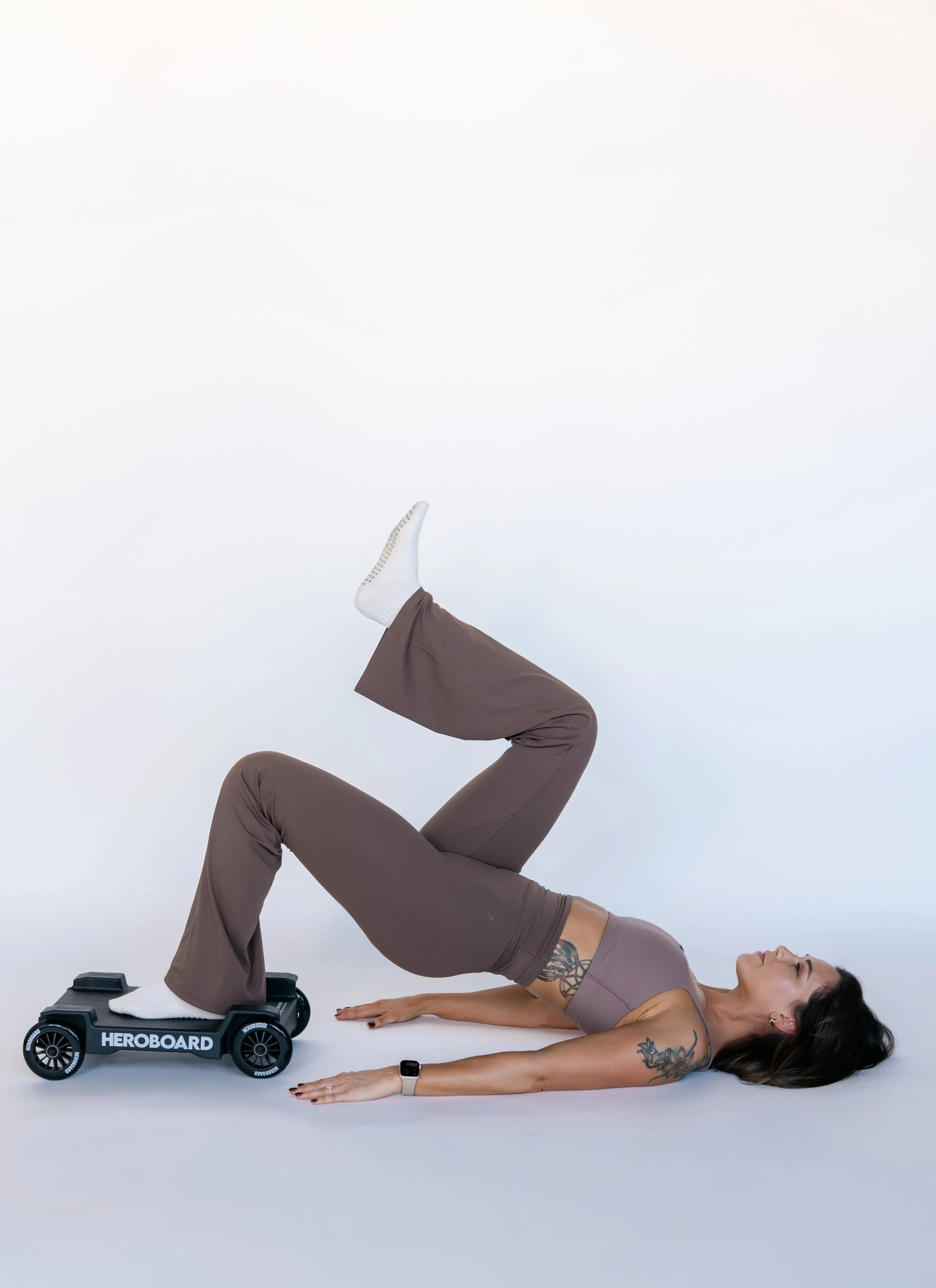 A woman exercising indoors on a white background, with one leg elevated on a skateboard labeled 'HEROBARD' and the other leg raised in a bent position. She is lying on her back with her arms extended forward, wearing athletic clothing and a smartwatch, demonstrating a fitness pose.