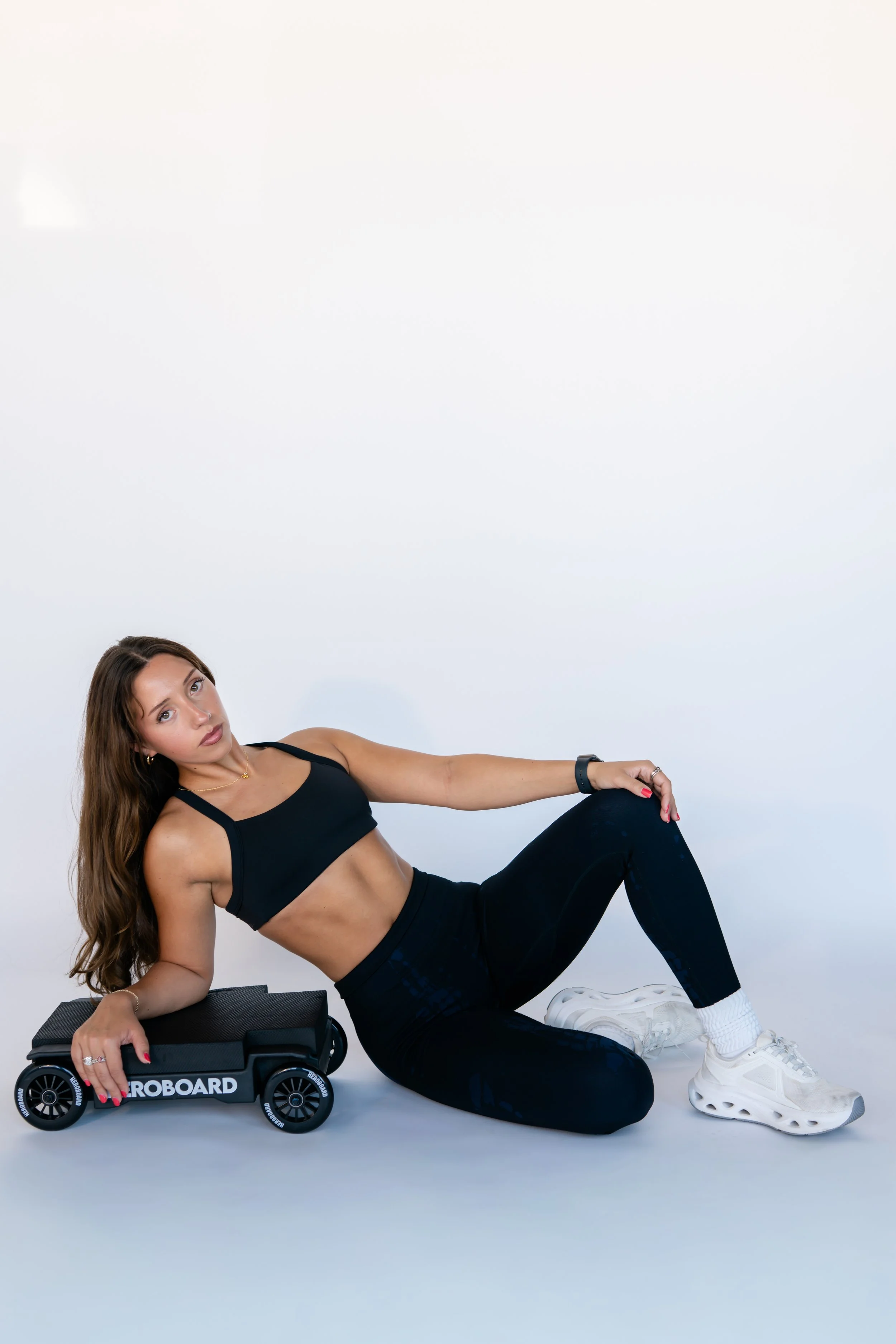 A woman with long brown hair wearing a black sports bra and leggings, sitting on the floor and leaning on her left arm, with her right arm resting on a skateboard labeled 'ICE BOARD', against a plain white background.