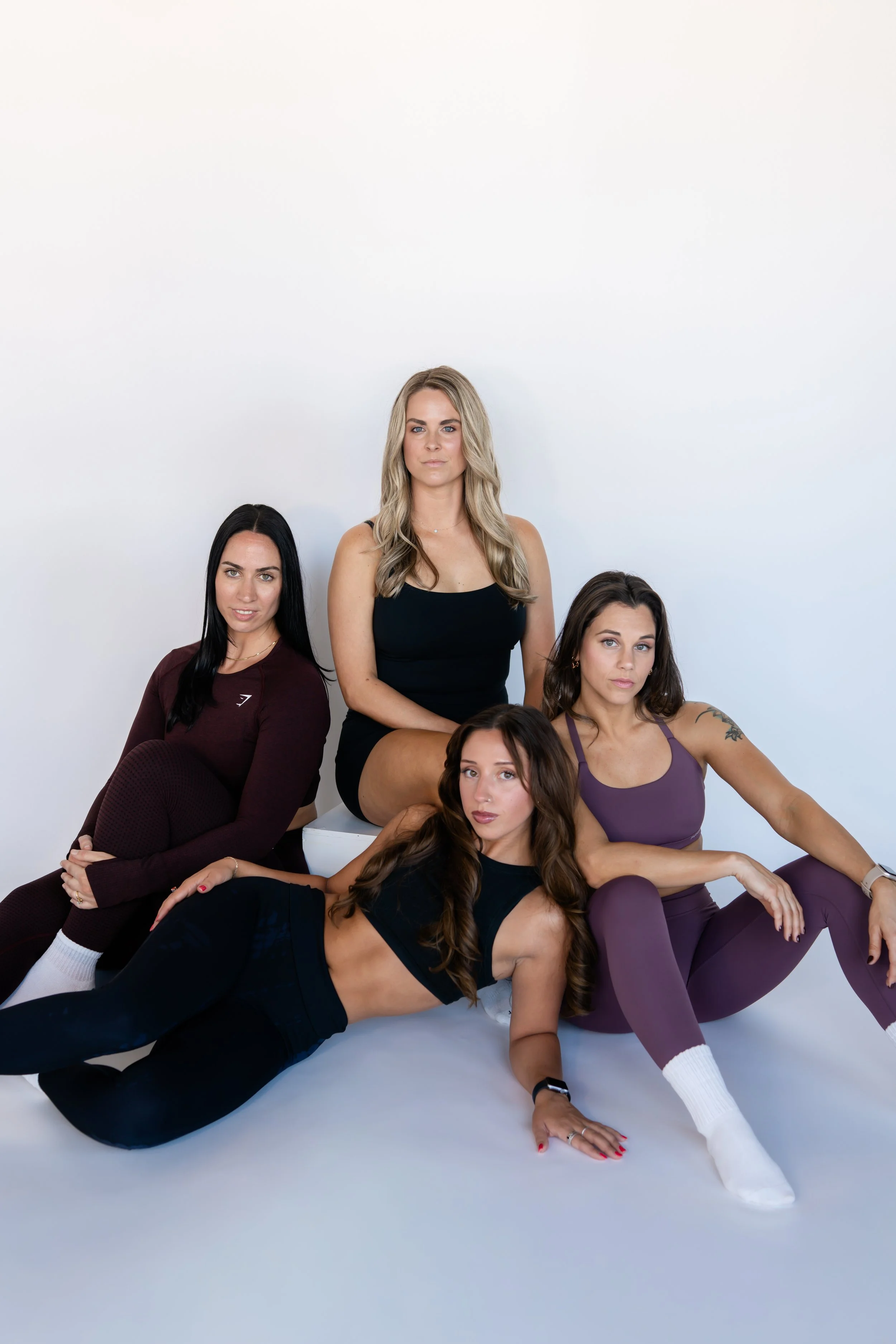 Group of five women with varying hair colors and styles, posing together against a plain white background. Some are sitting, some are standing, and they are dressed in casual athletic clothing.