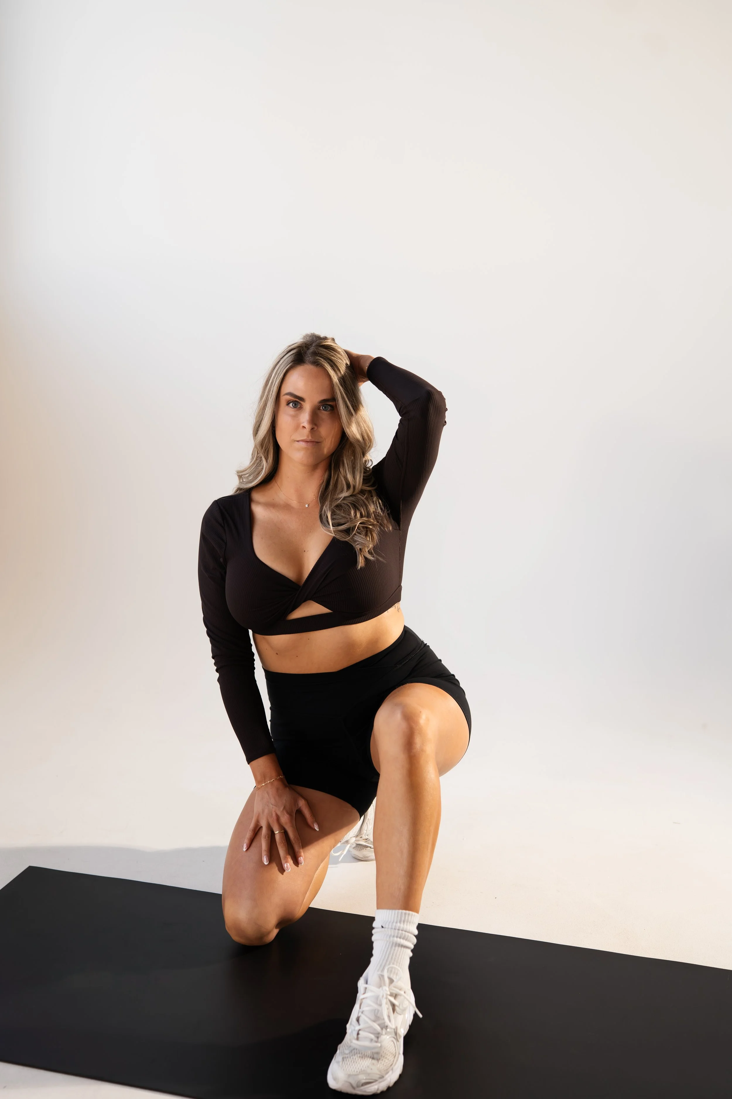 A woman with long hair wearing a black crop top, black shorts, white sneakers, and white socks is posing on a black yoga mat against a white background. She is kneeling on one knee, with one hand on her knee and the other hand behind her head, looking directly at the camera.