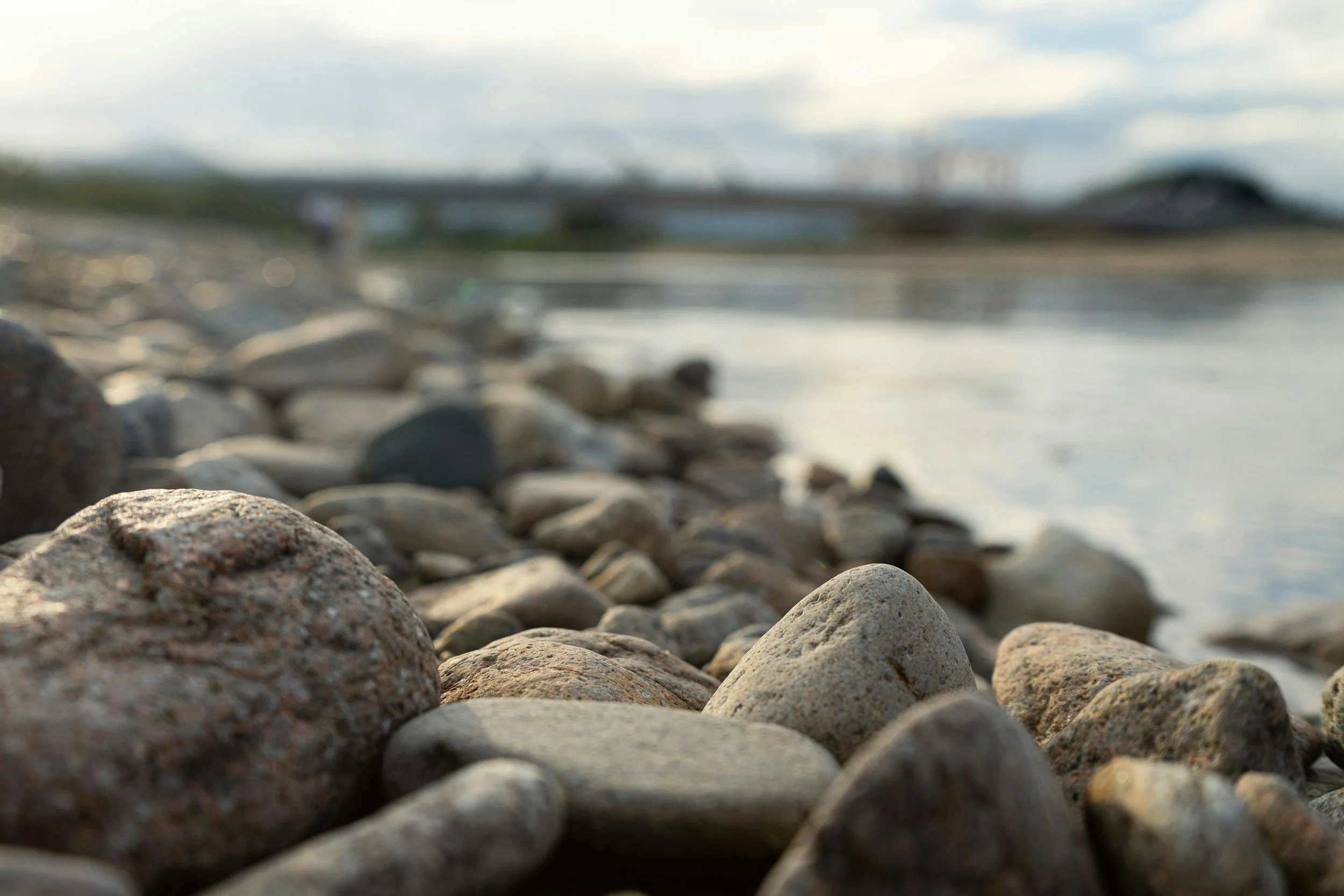 Close-up of smooth rocks and pebbles on the shore of a body of water, with a blurred background of a bridge and sky.