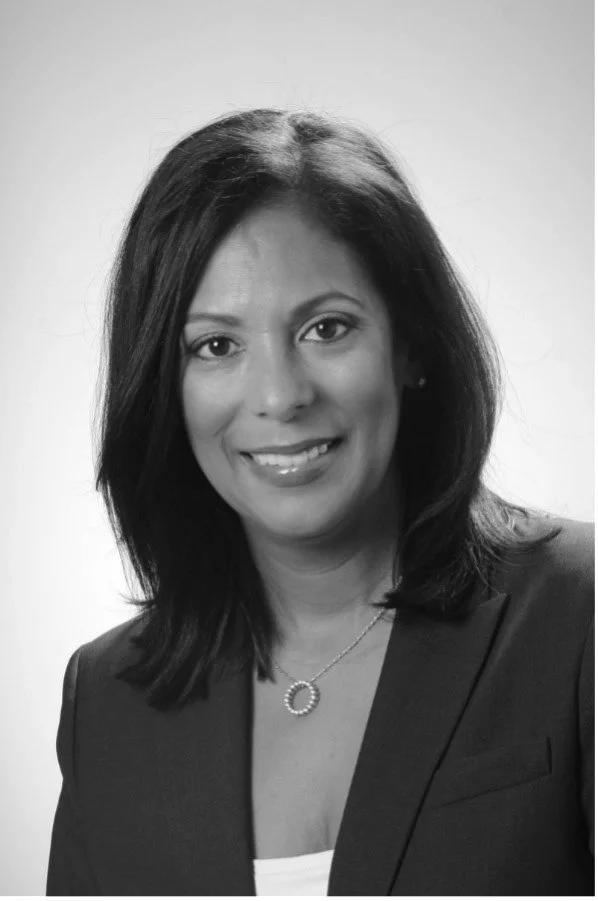 Black and white portrait of a woman with medium-length hair, wearing a blazer and necklace, smiling at the camera.
