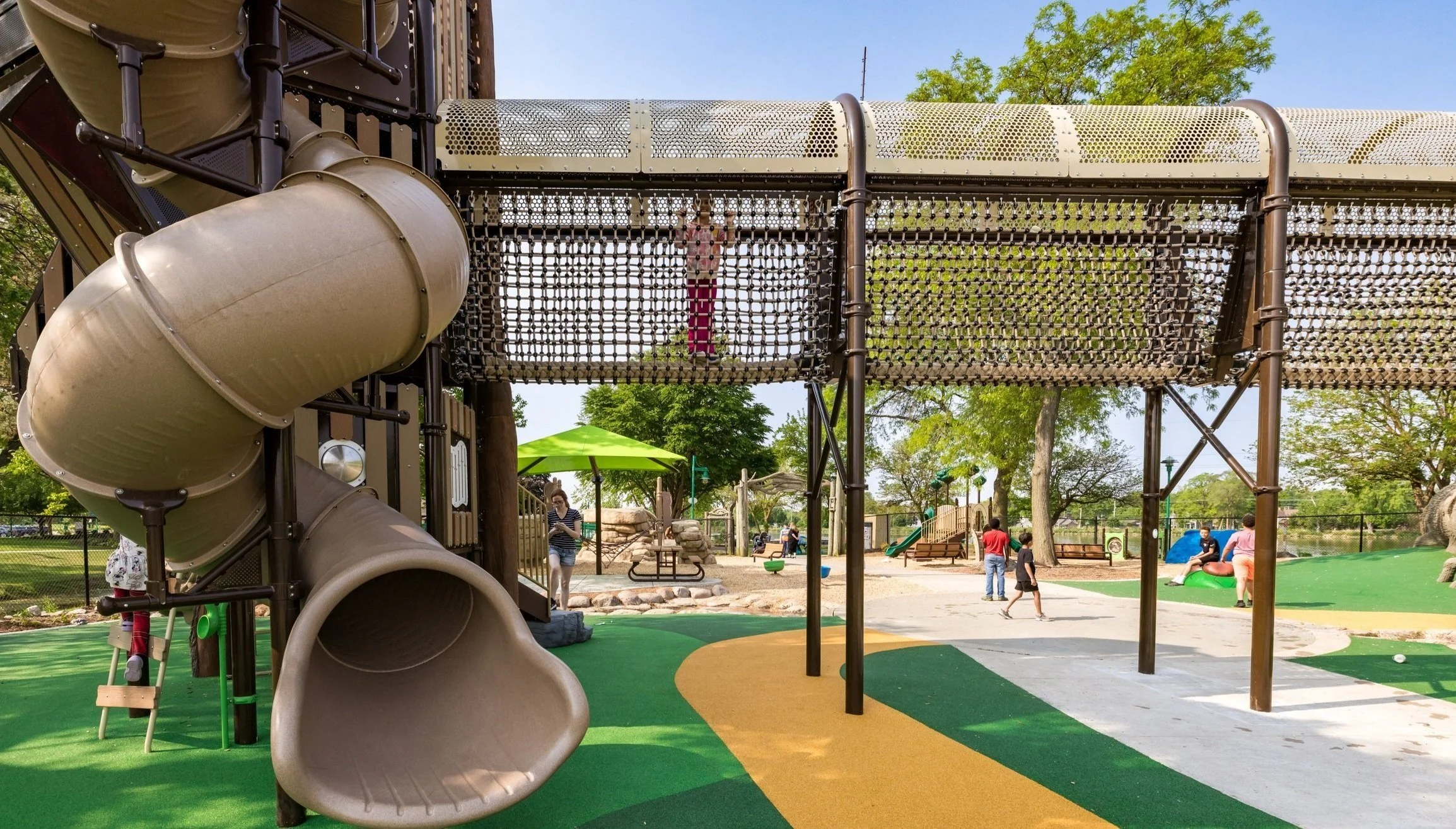 Children playing on the playground with slides, climbing structures, and trees in the background on a sunny day.