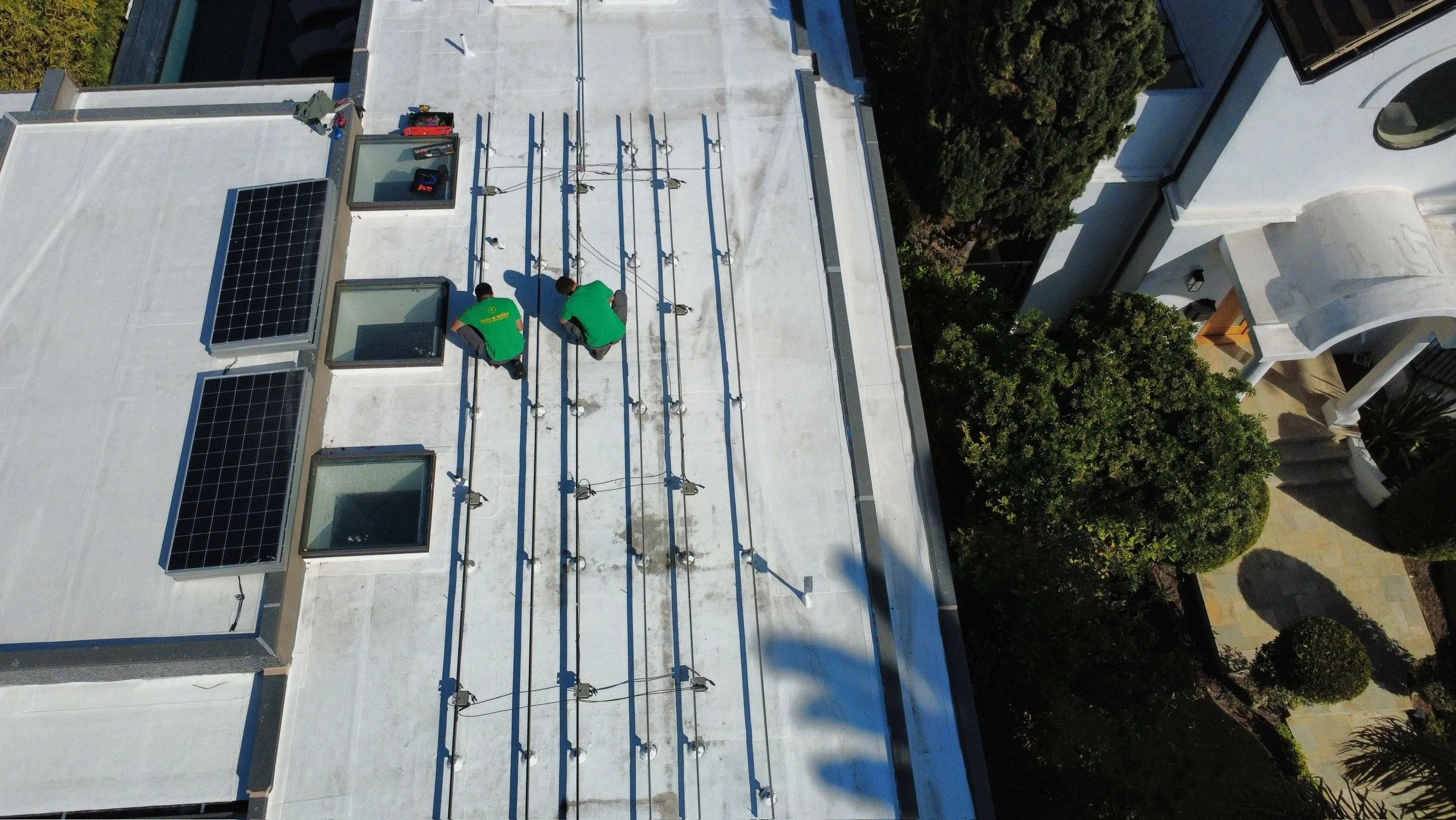 Two workers installing or repairing solar panels on the roof of a building during daytime, with green trees and a white curved structure nearby.