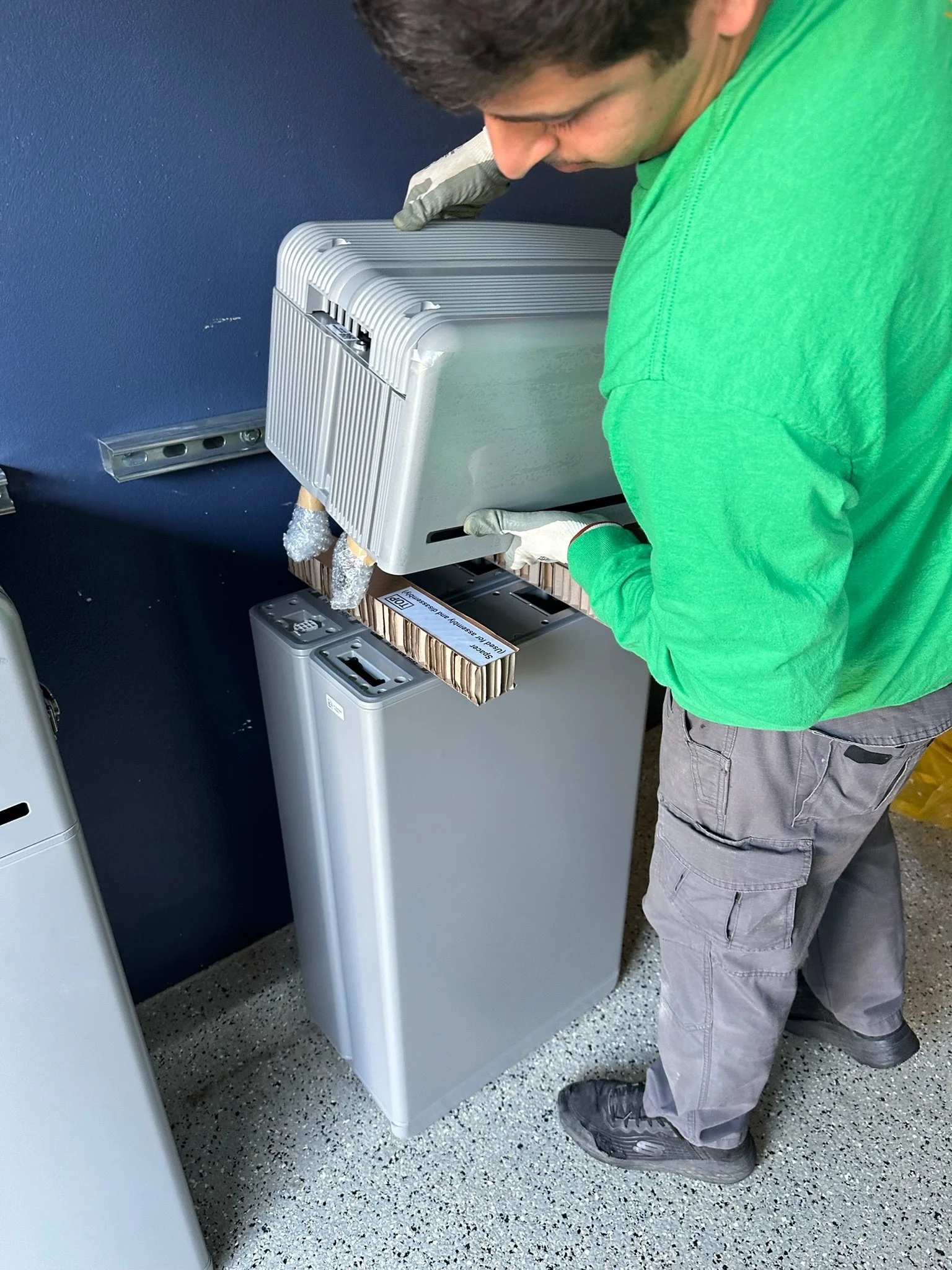 A person installing or repairing a gray sidebar shredder in an office or workspace, wearing gloves, green shirt, gray pants, and black shoes.