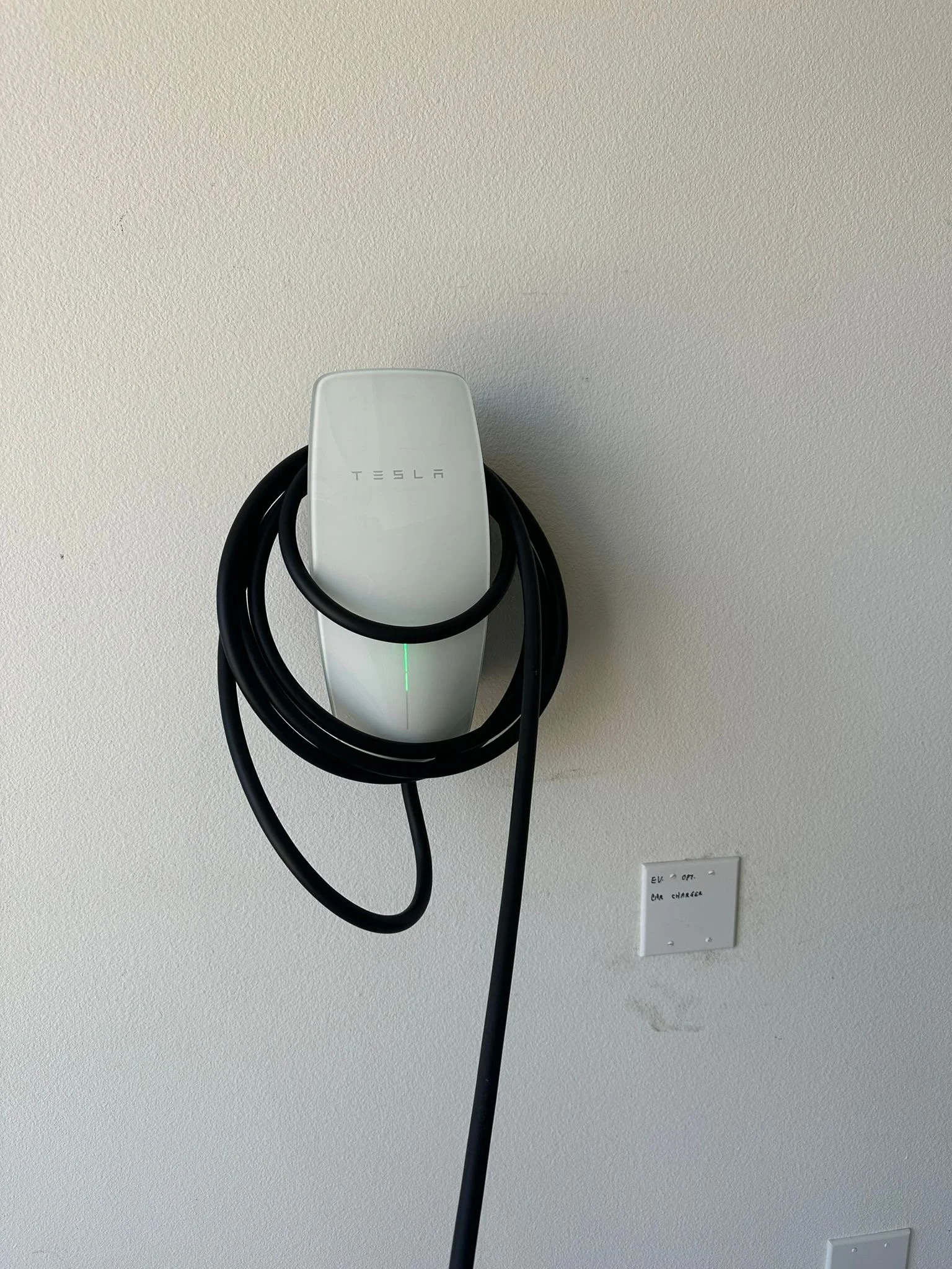 Tesla electric vehicle charger mounted on a ceiling with a black charging cable hanging from it, and an electrical outlet on the ceiling nearby.