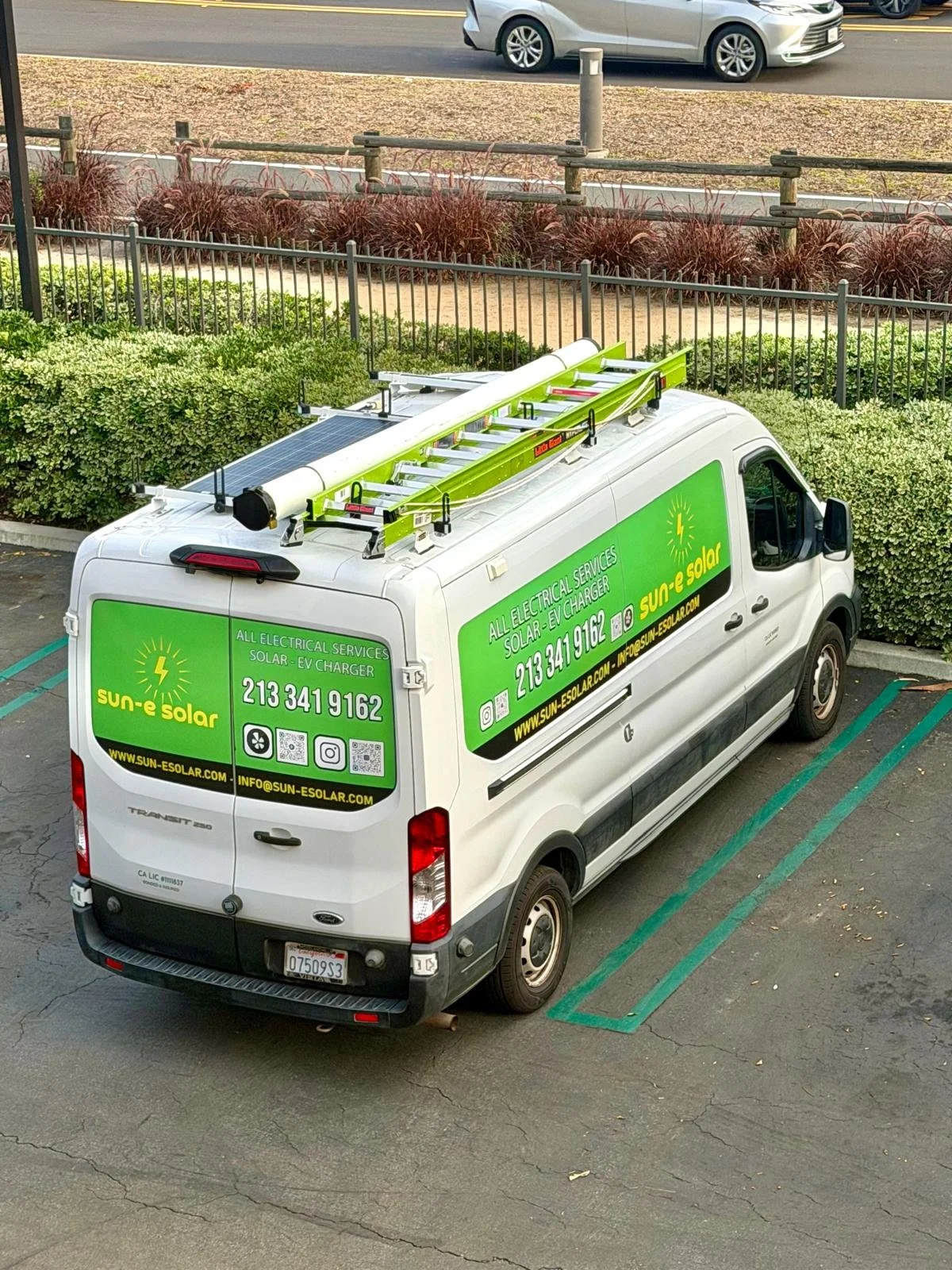 A white work van with green and yellow branding for Sun-e Solar, parked in a parking lot. The van has a ladder and solar panel on its roof.