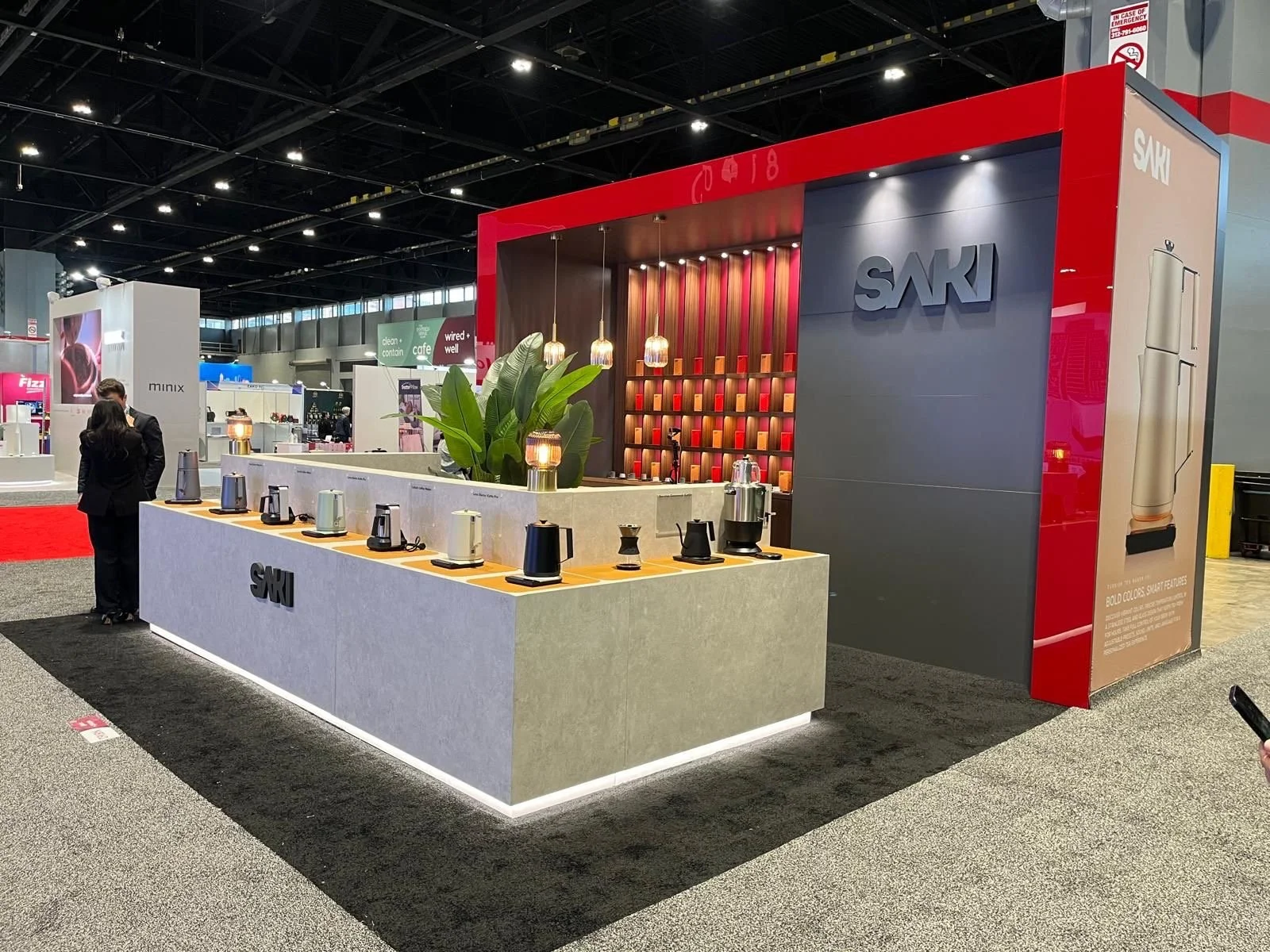 A trade show booth for SAKI, displaying various electric kettles on a countertop with a large green plant and small decorative lamps. The booth has a modern design with a red and gray color scheme, featuring the SAKI logo and a display of orange and red candles on a backlit wall. In the background, other booths and attendees are visible.