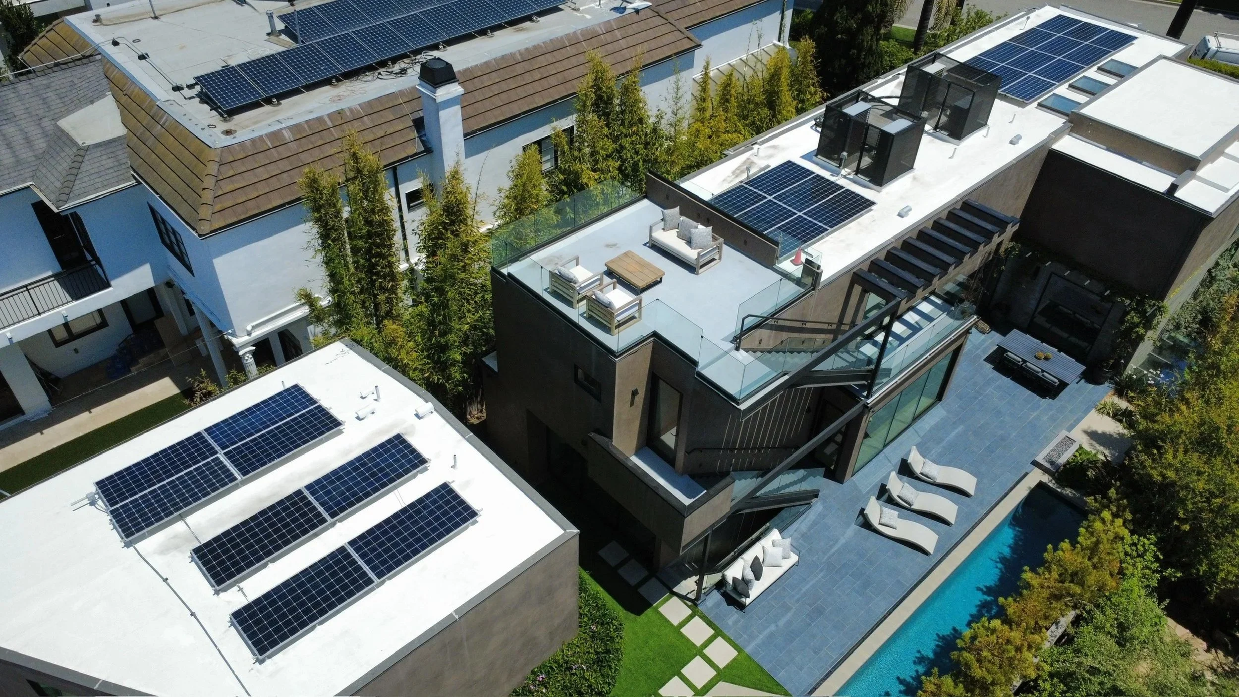 Aerial view of modern residential rooftops featuring solar panels, rooftop terrace with outdoor furniture, and a swimming pool surrounded by trees and landscaping.