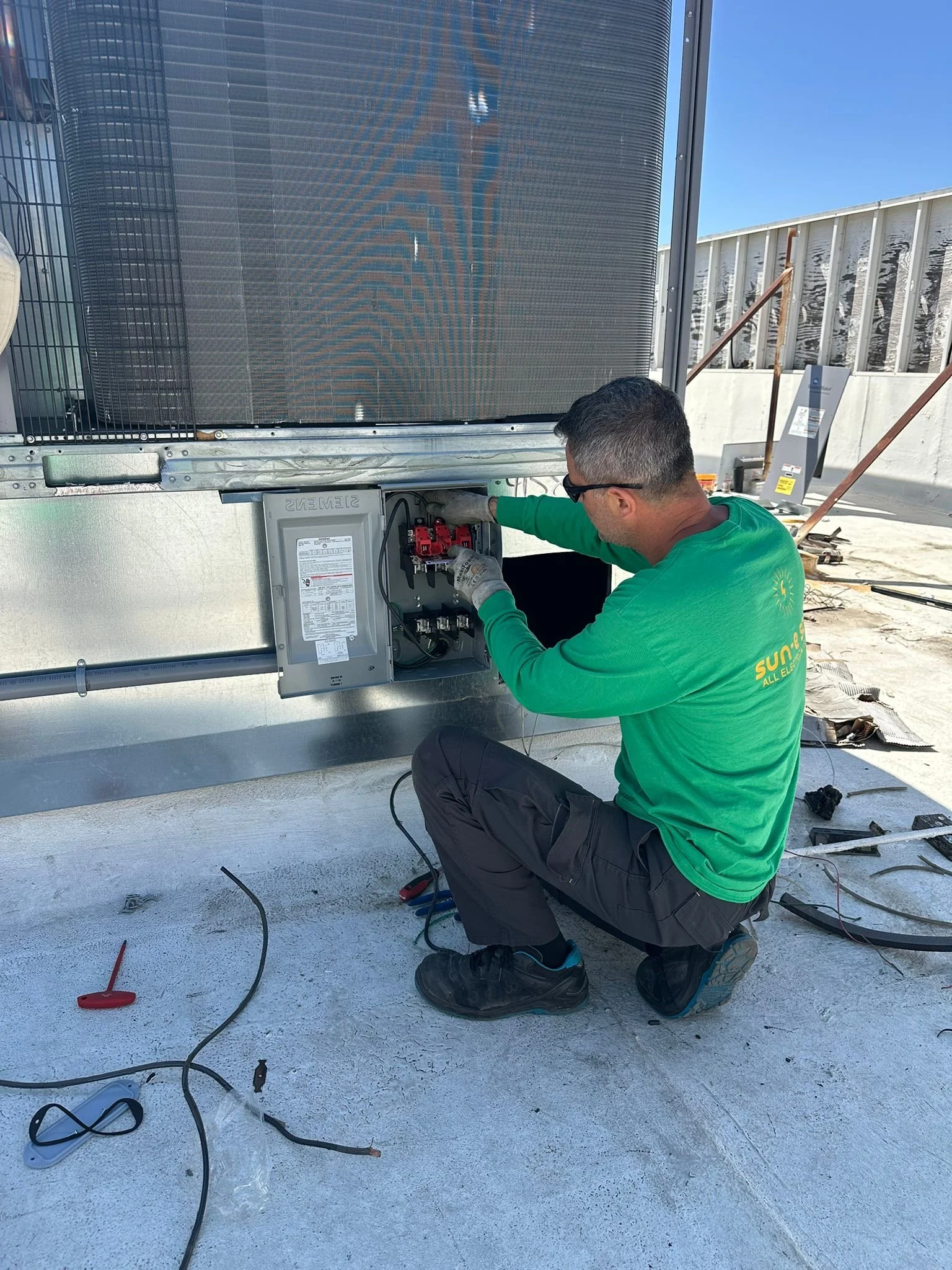 A technician working on an HVAC unit on a rooftop, connecting electrical components, wearing safety gear and a green shirt.