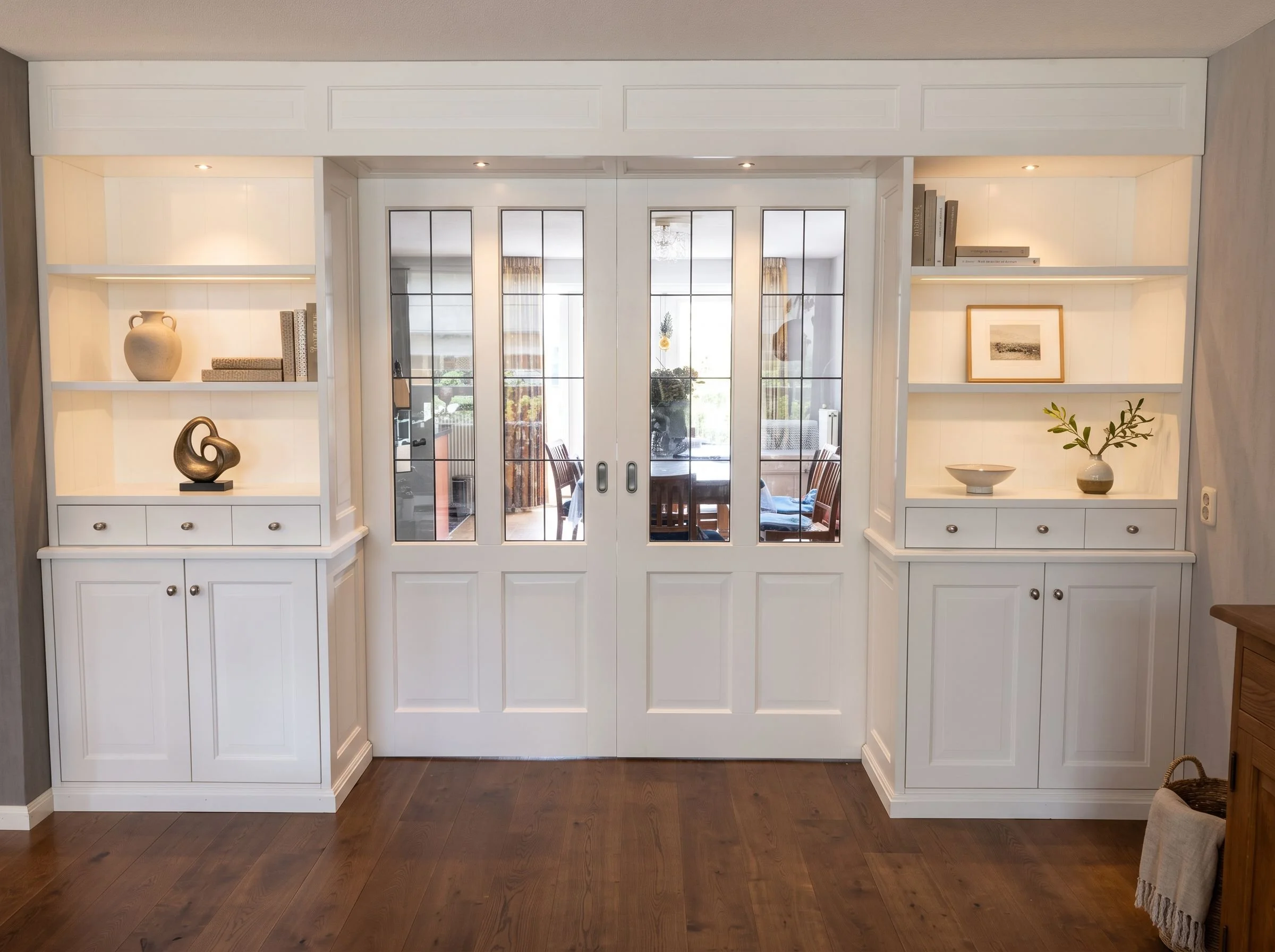 Witte ingebouwde kast met glazen schuifdeuren, decoratieve objecten en boeken binnen, in een kamer met houten vloeren.