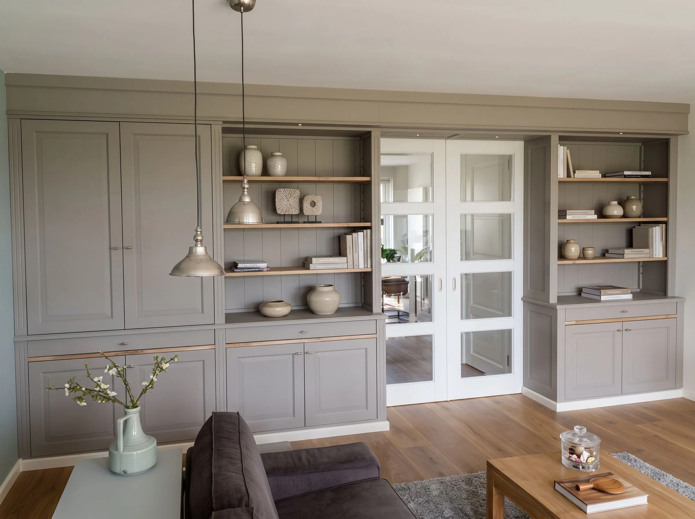 Woonkamer met grijze houten boekenkast, witte muren, houten vloer, salontafel, stoffen fauteuil, vaas met bloemen, en decoratieve vazen en boeken op de planken.
