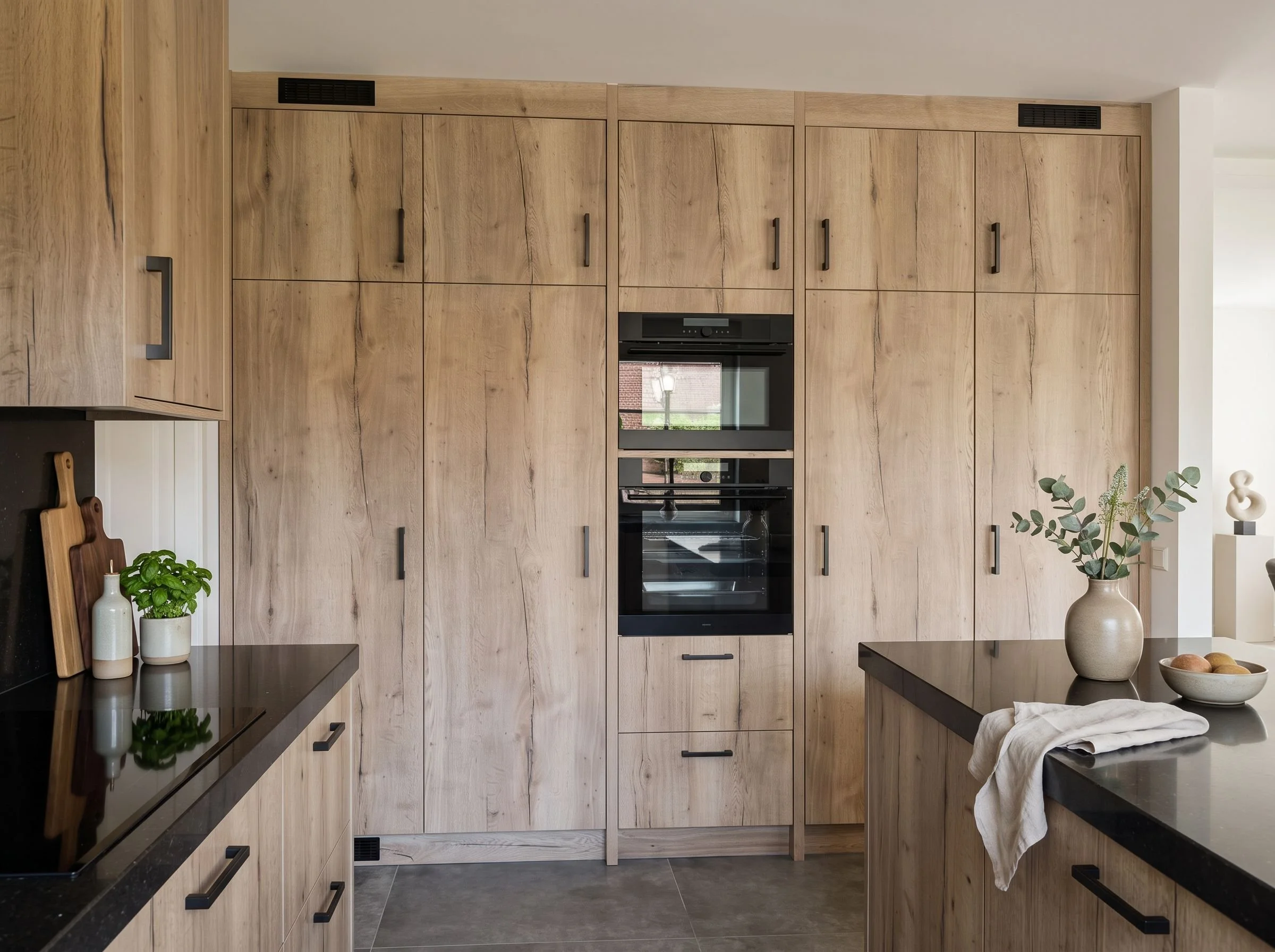 Een moderne keuken met houten kasten, een ingebouwde oven en magnetron, en zwart granieten werkbladen. Op het werkblad staan drie witte vazen met groen blad en een snijplank.