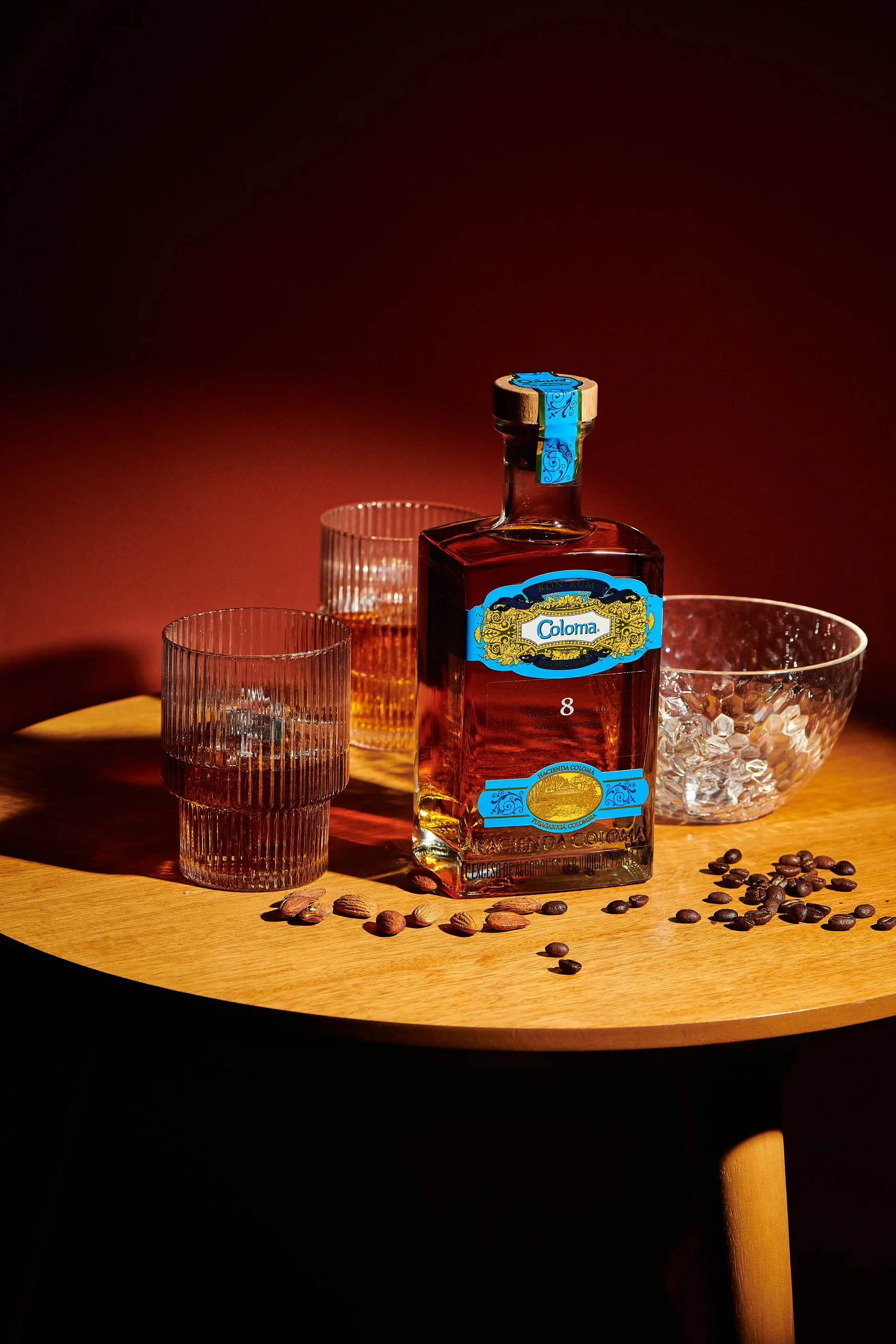 Botella de ron Coloma sobre mesa de madera, acompañado de tres vasos de diferentes estilos y una bowl con hielo, con granos de café dispersos en la mesa, fondo oscuro con luz en un lado