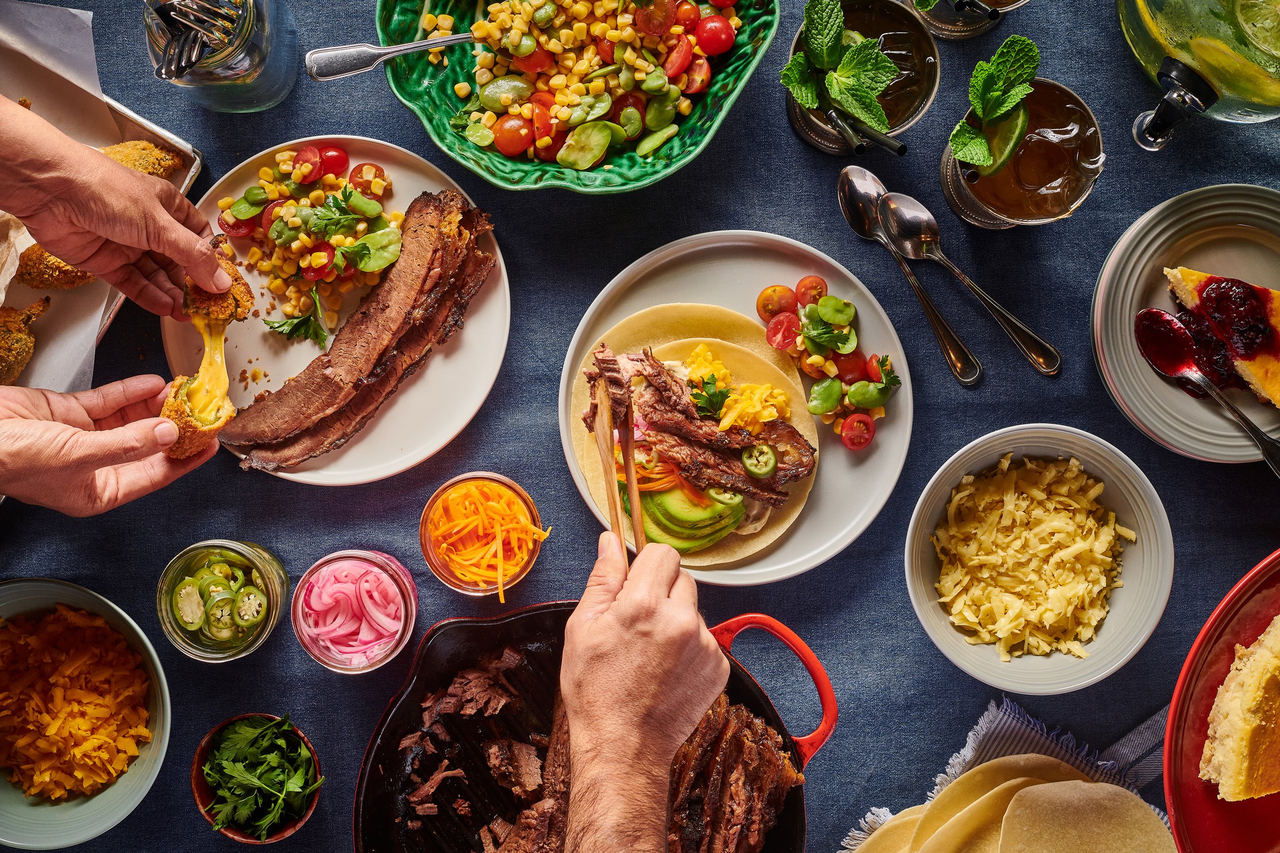 Mesa con diferentes alimentos tradicionales mexicanos, como tacos, carnes, ensaladas, salsas, queso rallado, y bebidas, para compartir