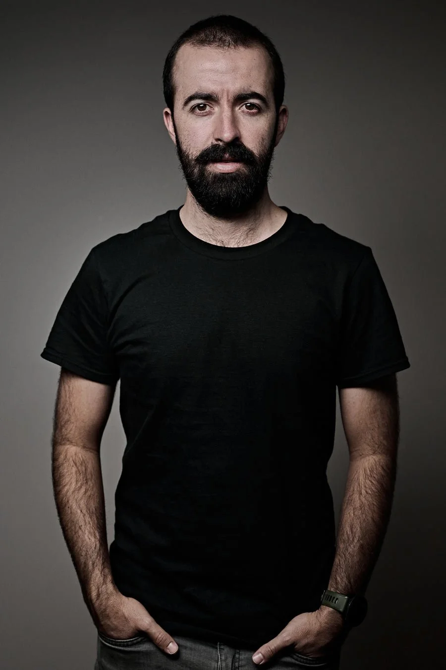 Hombre con barba, cabello corto y camiseta negra, con fondo gris oscuro.