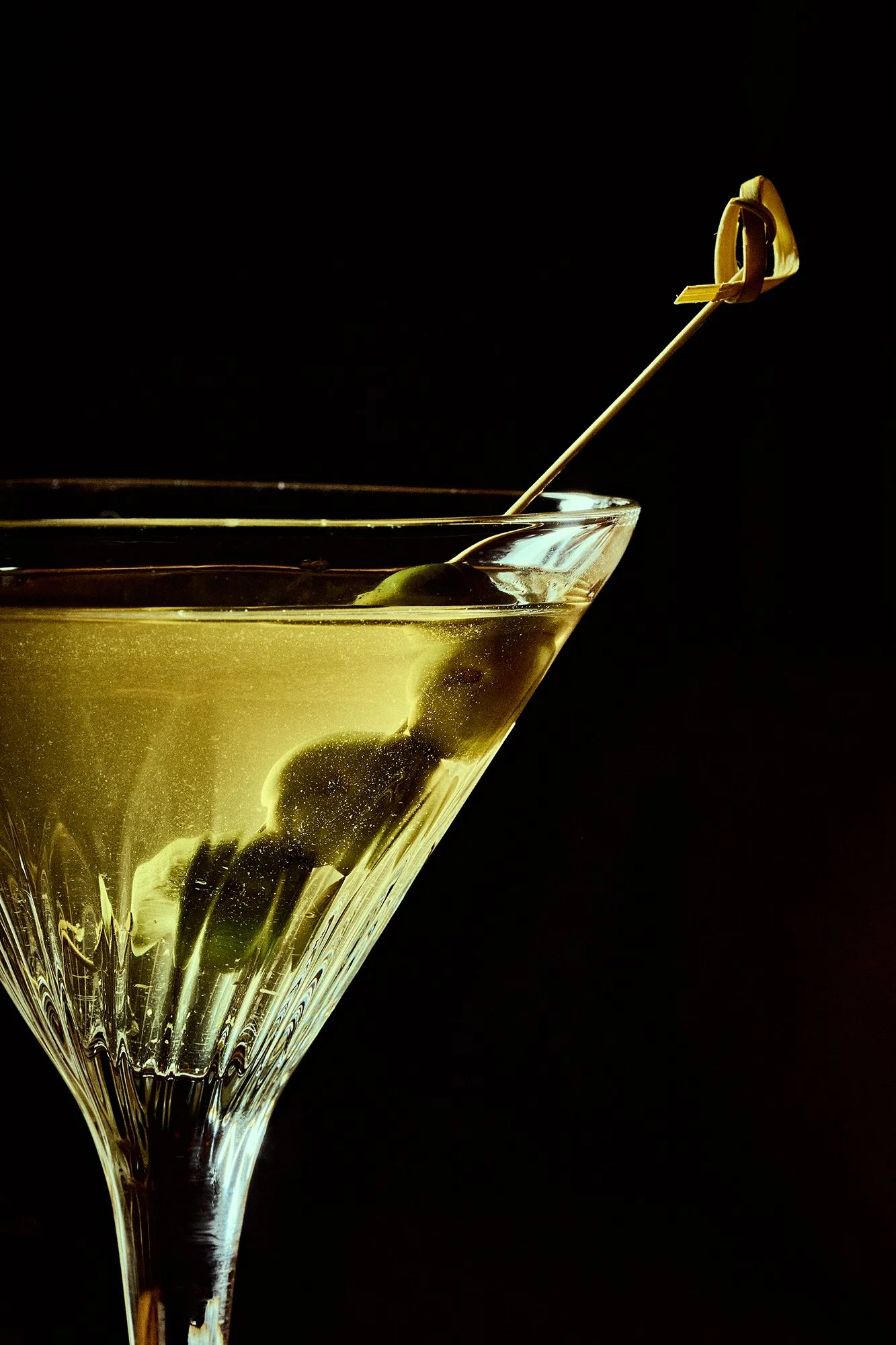 Copa de cóctel con aceitunas verdes y una aceitera dentro, decorada con un agitador de cóctel en un fondo negro.