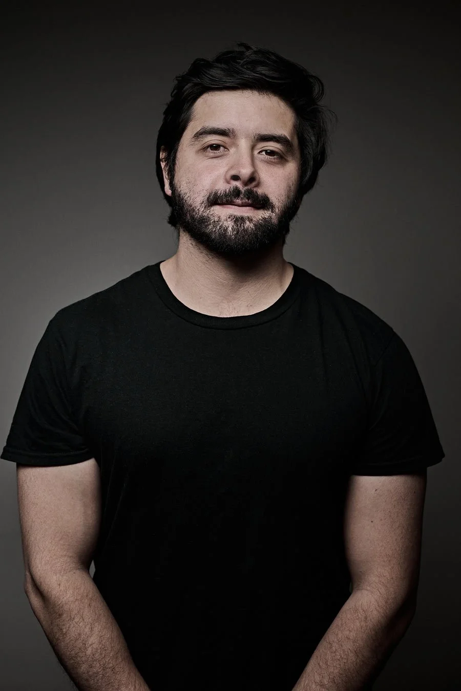 Retrato de un hombre con barba y cabello oscuro, usando una camiseta negra, con fondo gris oscuro.