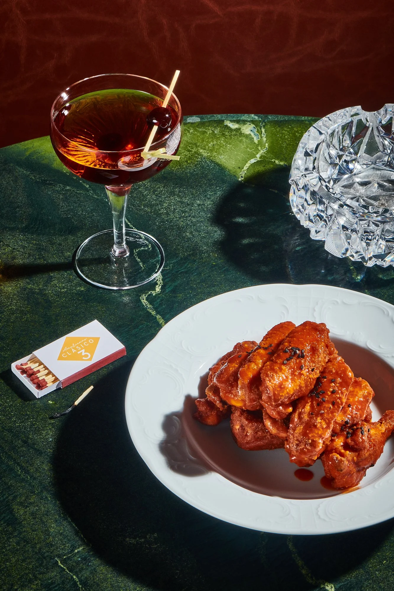 Un cóctel con cereza y una tapa de alitas de pollo fritas con salsa en un plato. También hay una caja de cerillas y un cenicero de vidrio en la mesa.