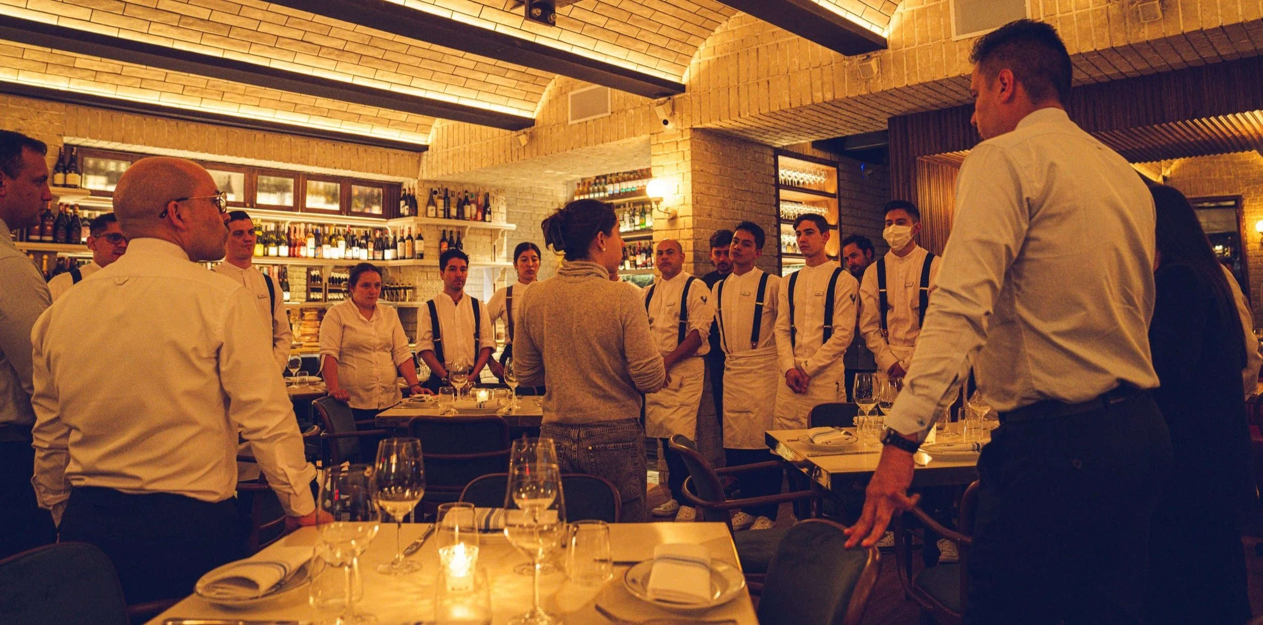 Grupo de camareros y una mujer en una reunión en un restaurante con decoración cálida y mesas con cristalería y manteles blancos.
