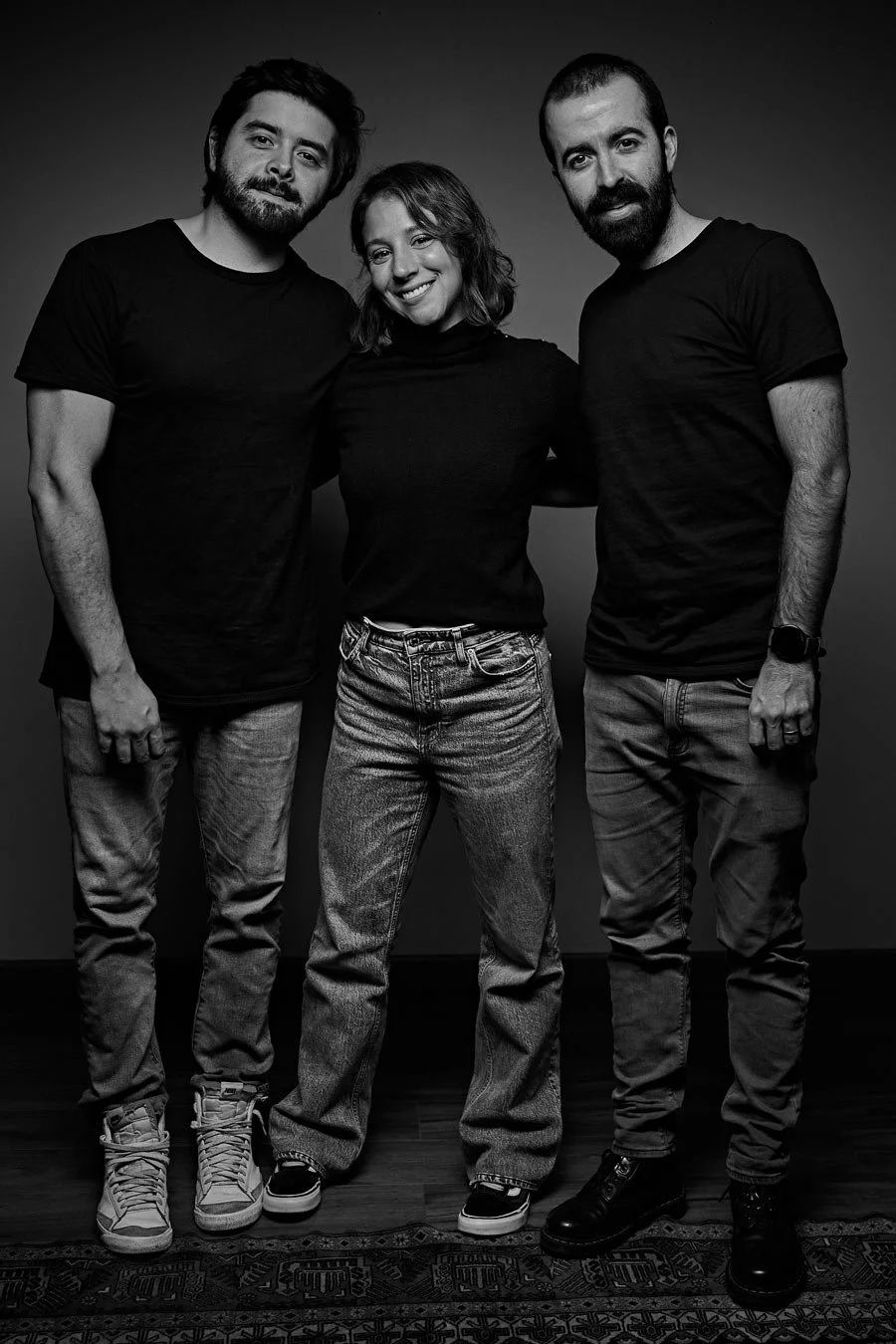 Tres personas, dos hombres y una mujer, posando juntos en un estudio con fondo gris, todos vestidos con camisetas negras y jeans, sonriendo en una foto en blanco y negro.