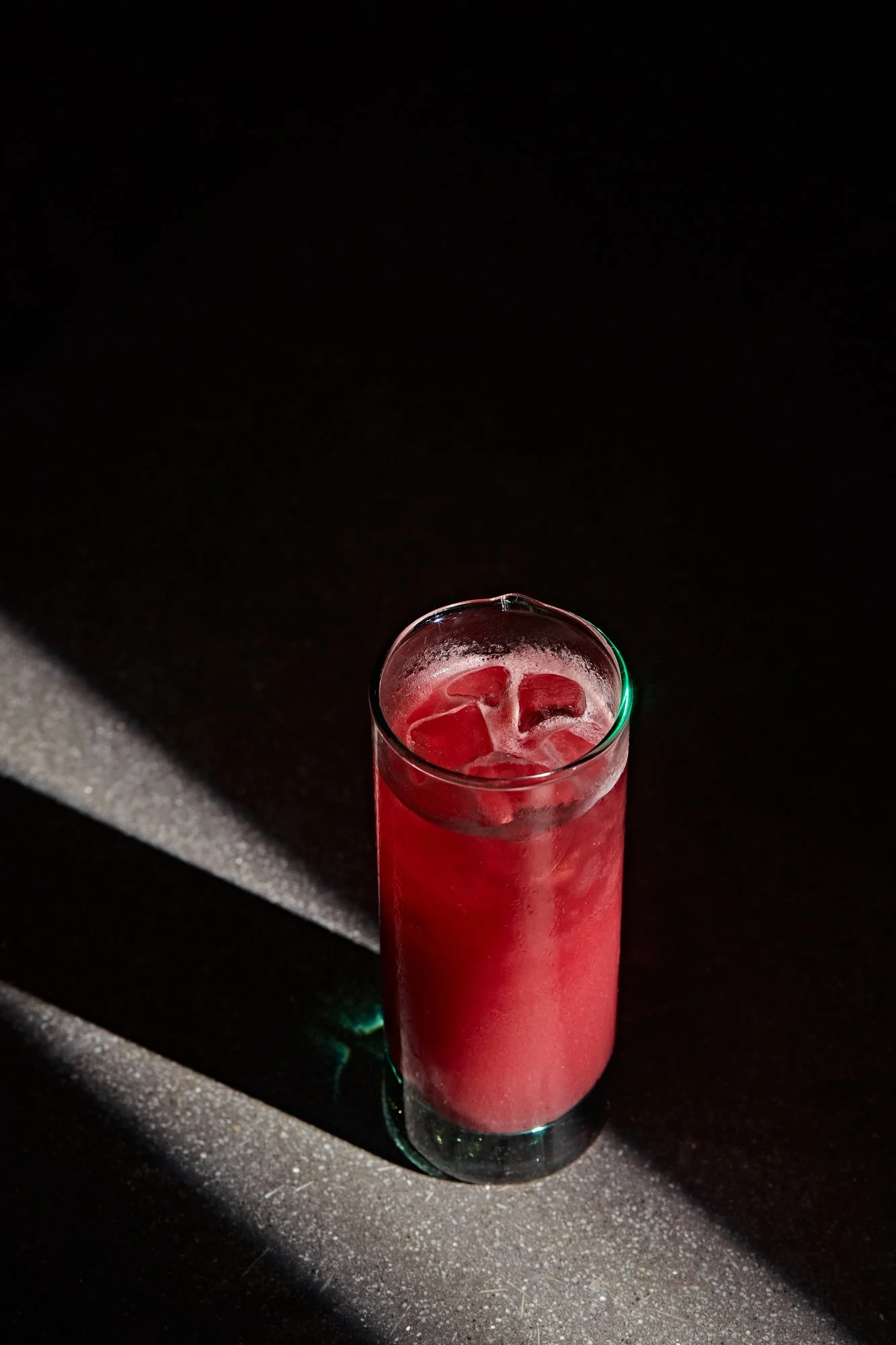 Vaso alto con bebida rosada y cubitos de hielo, en una mesa de superficie gris oscuro, sombreada por una luz que crea un contraste con el fondo negro.