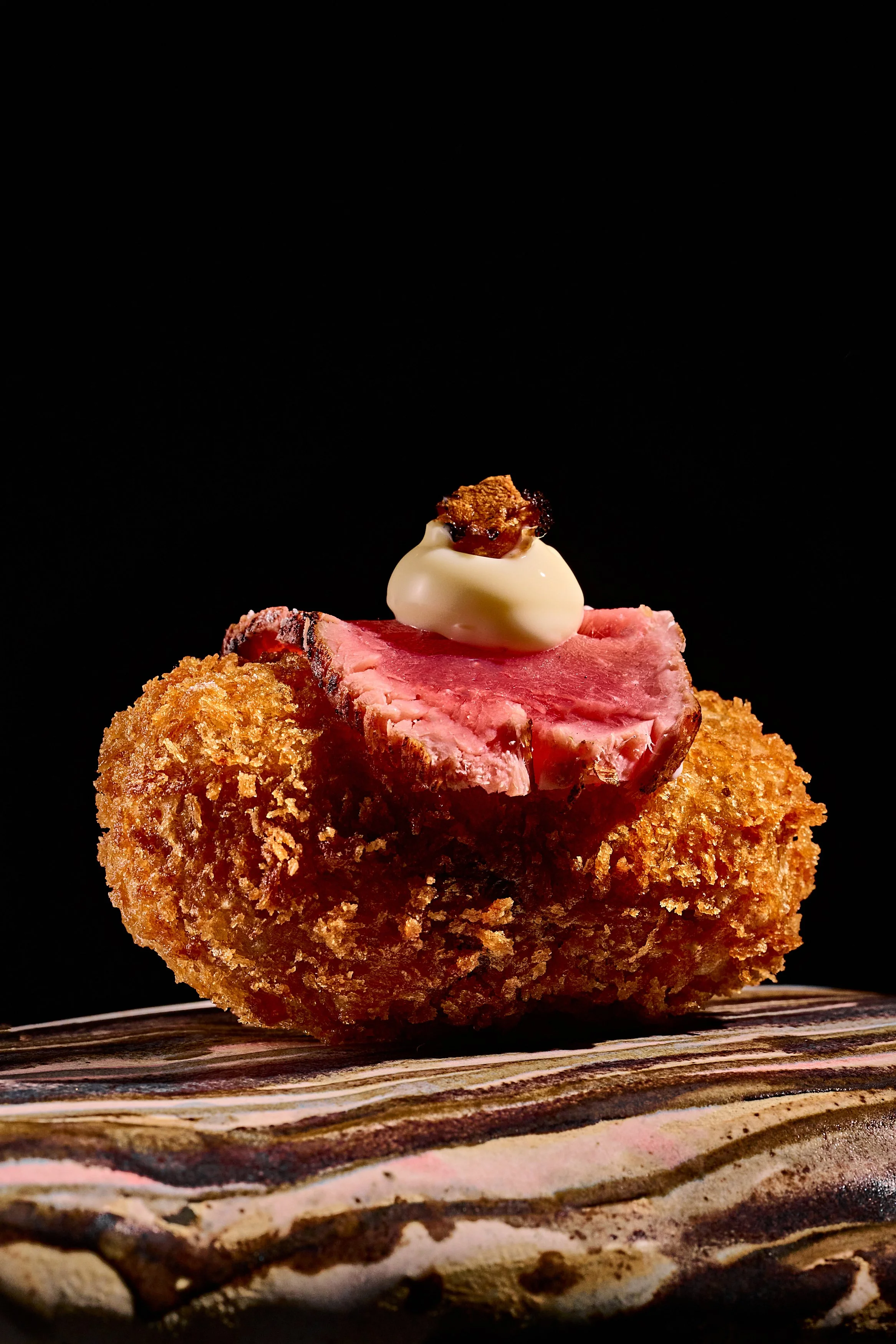 Croqueta de carne empanizada con trozo de carne, una bola de salsa blanca y reducción de carne sobre ella, en un fondo negro