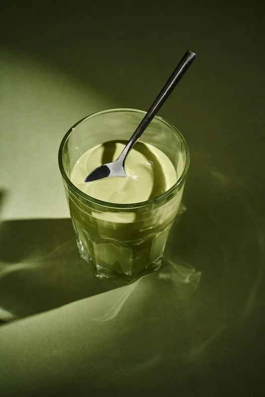 Vaso con una bebida yogurt de matcha y una cuchara dentro, sobre una superficie con sombra.