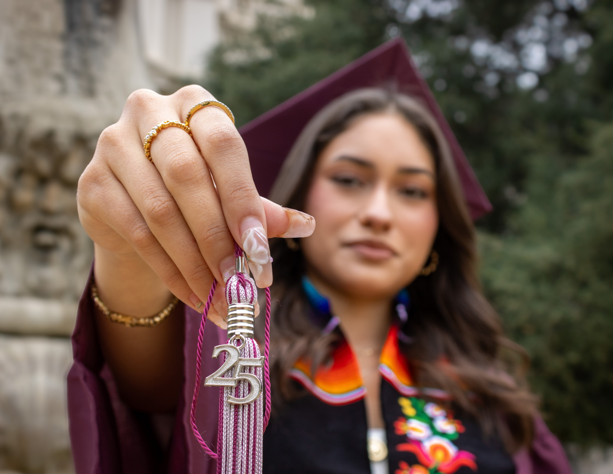 Close-up of a woman's hand holding a pink lanyard with a metallic number '25' and a red string, with her face slightly blurred in the background, wearing a maroon graduation cap and a colorful traditional outfit, outdoors with greenery.