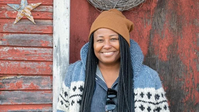 Krista Scruggs smiling in front of a rustic red wooden wall with a wreath and a star decoration, wearing a brown beanie, a cozy blue and white knit sweater, and braids.