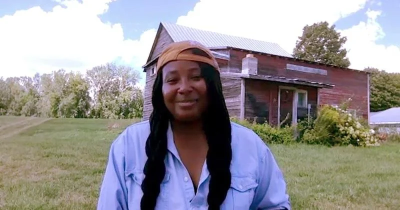krista scruggs with long braids wearing a backward cap and denim shirt, standing in front of a rustic barn in a grassy field