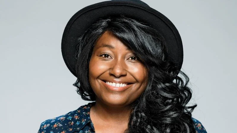 krista scruggs smiling wearing a black hat with long wavy hair and a floral-patterned shirt