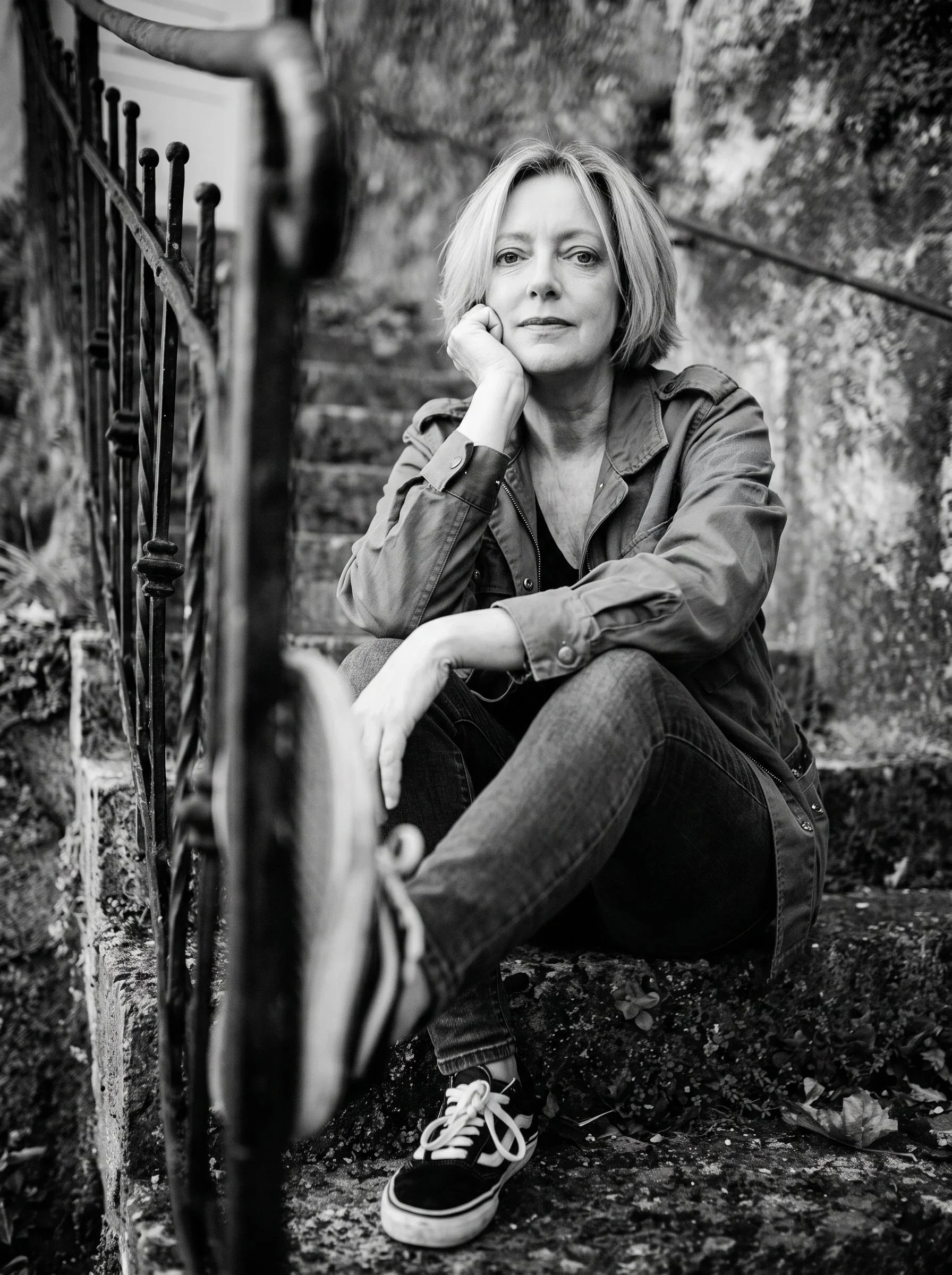 Angelique, psychic medium and intuitive guide, sitting on weathered stone steps beside an iron railing in a black and white self portrait