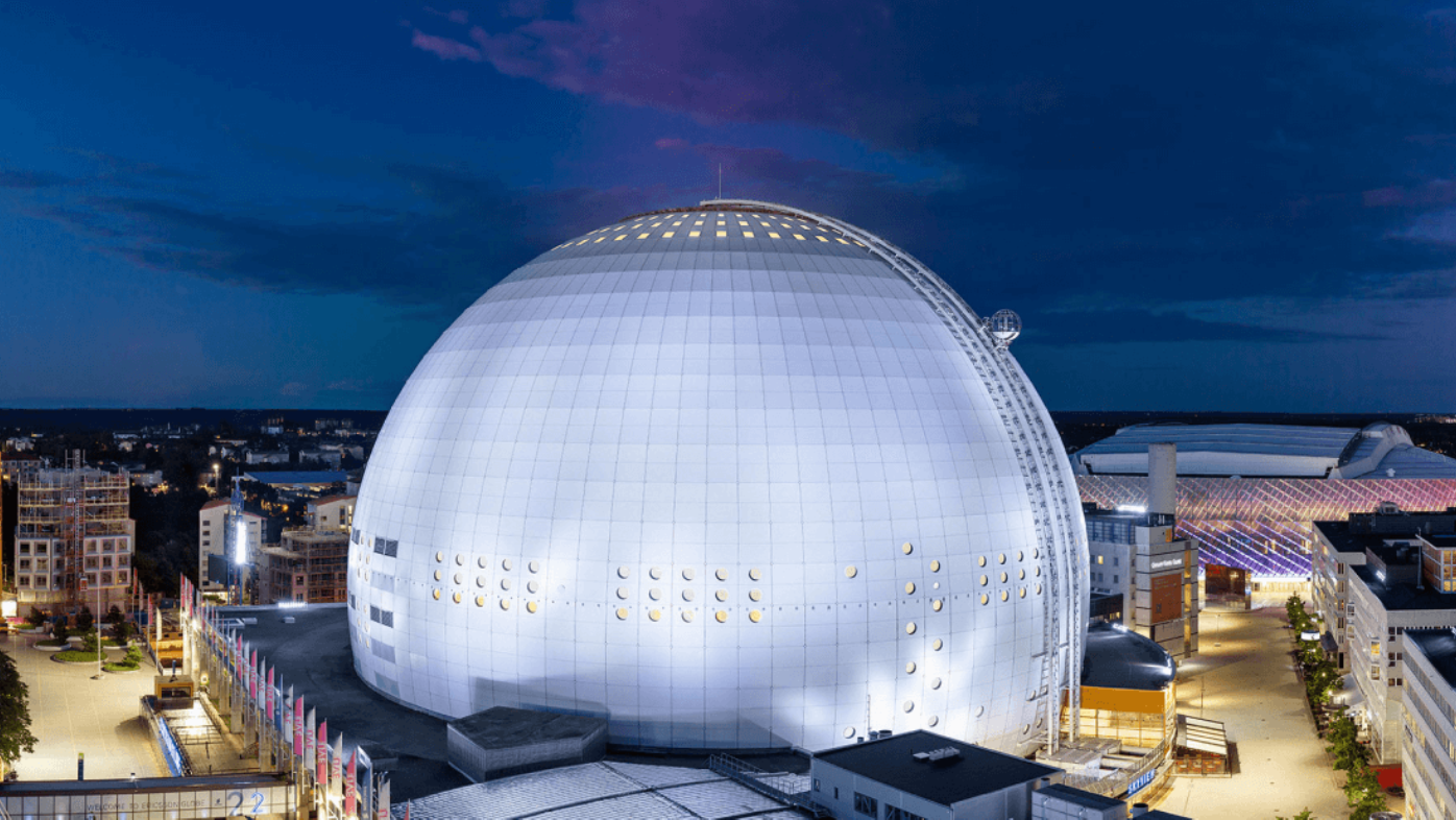 En stor, sfärisk byggnad med en modern design, belägen i en stad på kvällen. Byggnaden är belägen bland andra byggnader och omges av gatubelysning och byggnader.