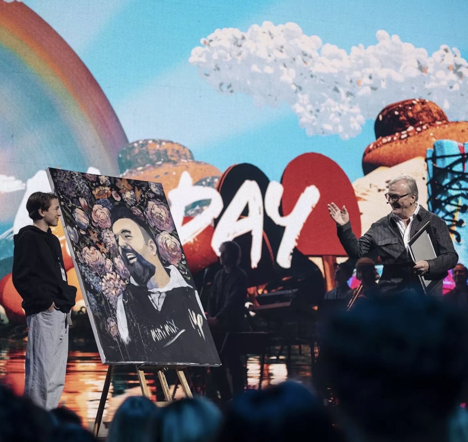 En person håller ett porträtt av en man med blommor runt omkring, medan en annan person talar med en mikrofon på en scen med en stor bakgrund som visar texten "2 DAY" och en regnbåge.