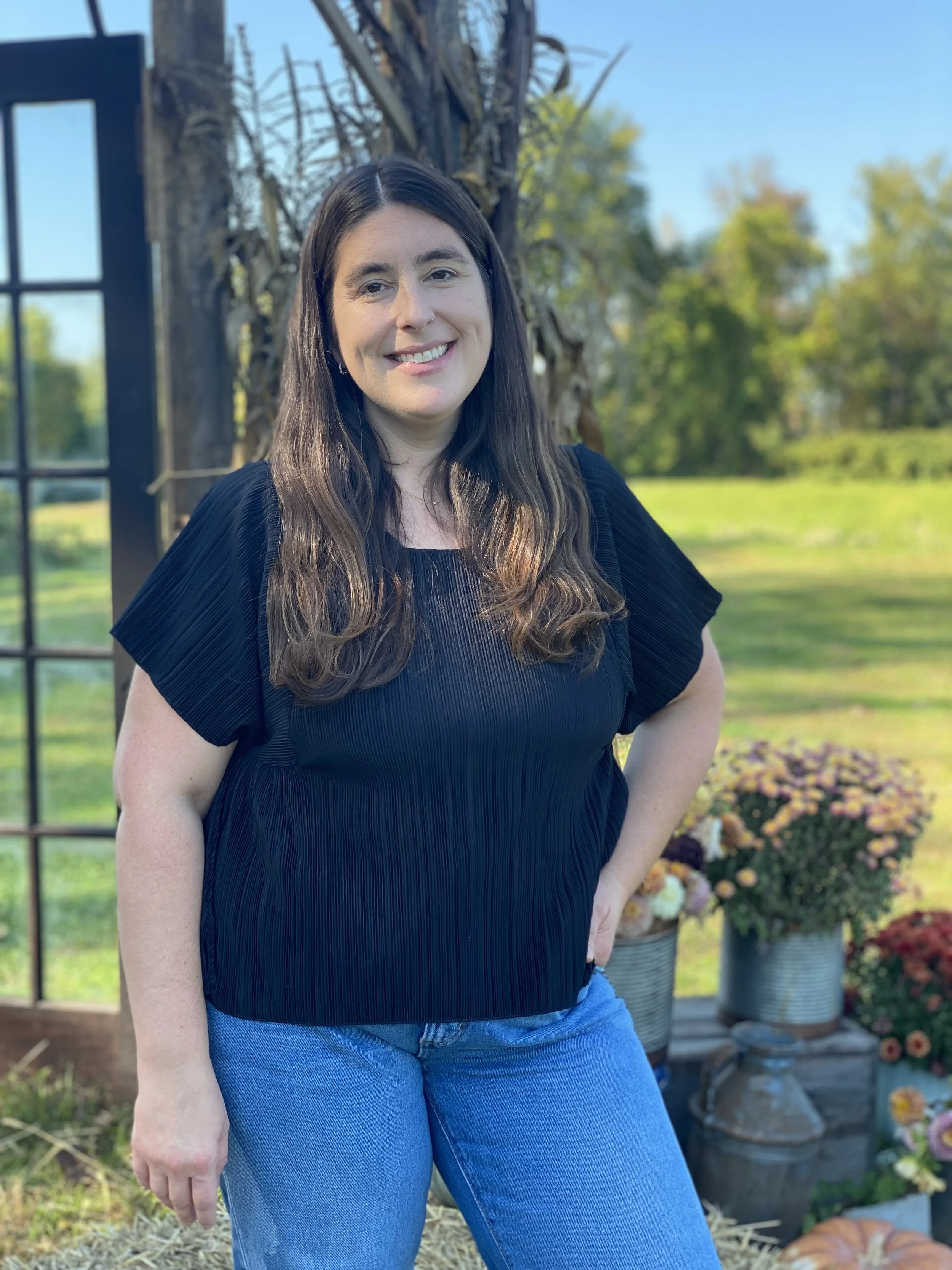A woman with long brown hair and fair skin standing outdoors smiling at the camera. She is wearing a black blouse and blue jeans. Behind her are potted flowers, a leafless tree, and a grassy background with trees and a blue sky.