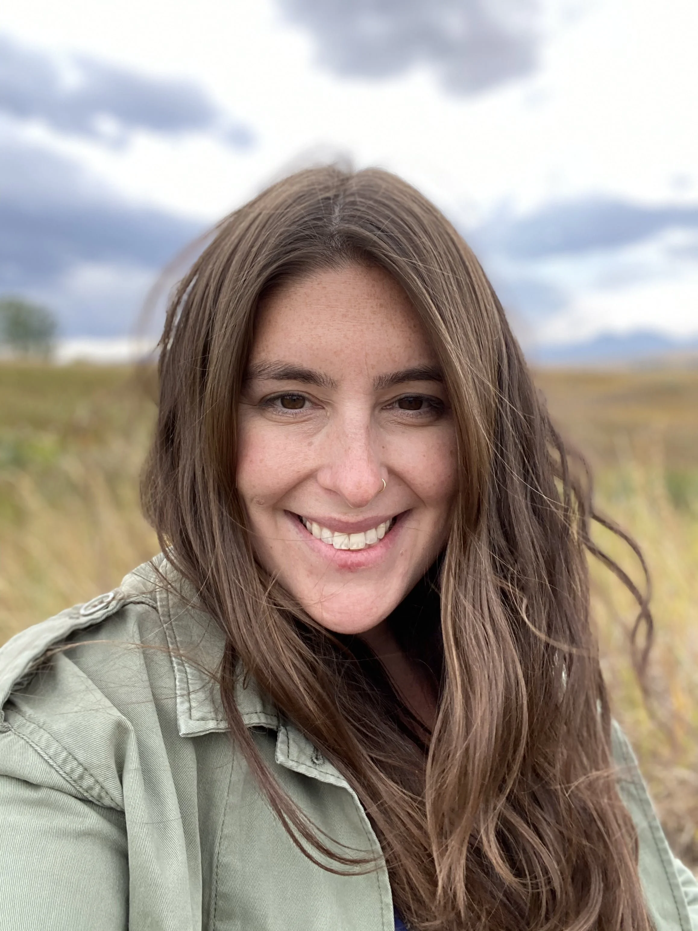 A woman with long brown hair, smiling, outdoors with a cloudy sky and field in the background.