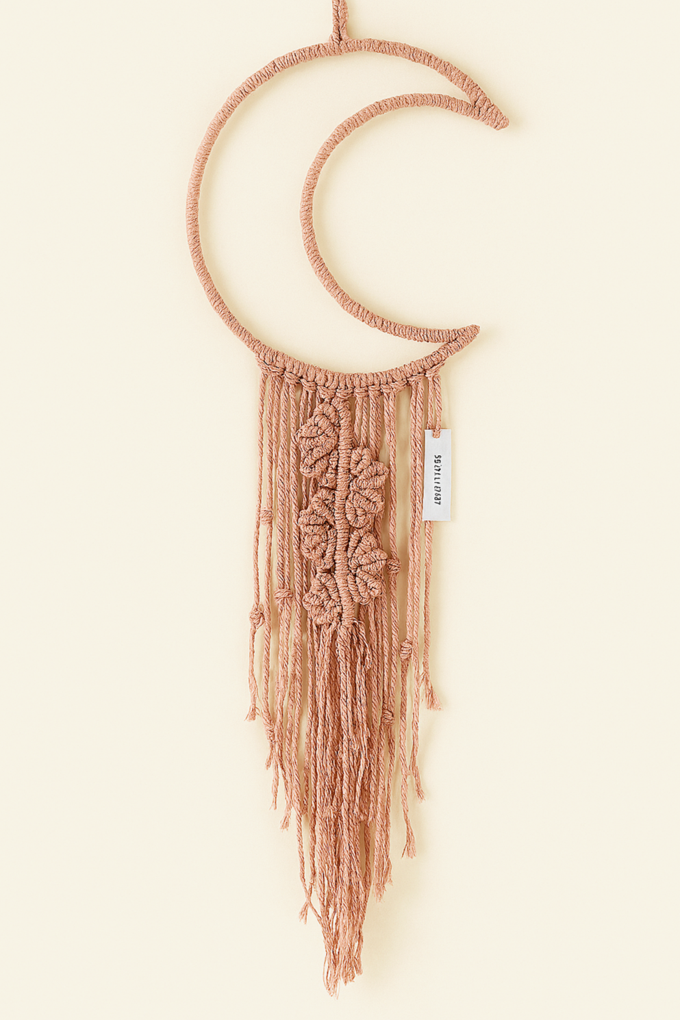 Boho macramé wall hanging shaped like a crescent moon in dusty pink, featuring floral-inspired knots and long fringe. Neutral backdrop, perfect for cozy home decor.