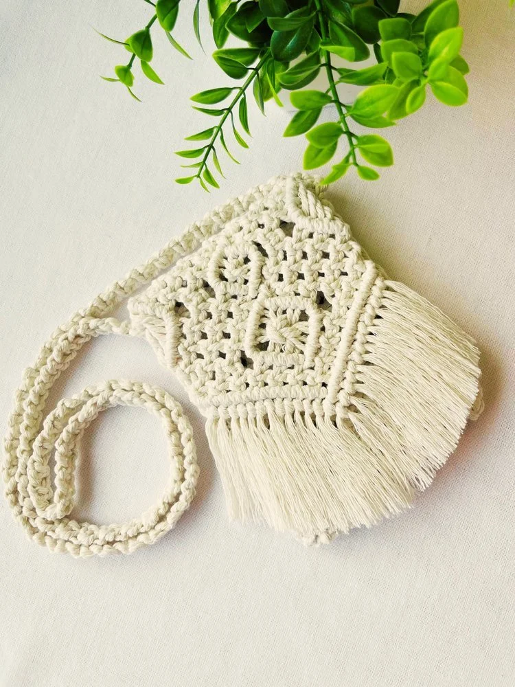 Handmade off-white macramé crossbody bag with fringe and intricate knot design, styled with green leaves – Samra Knots.