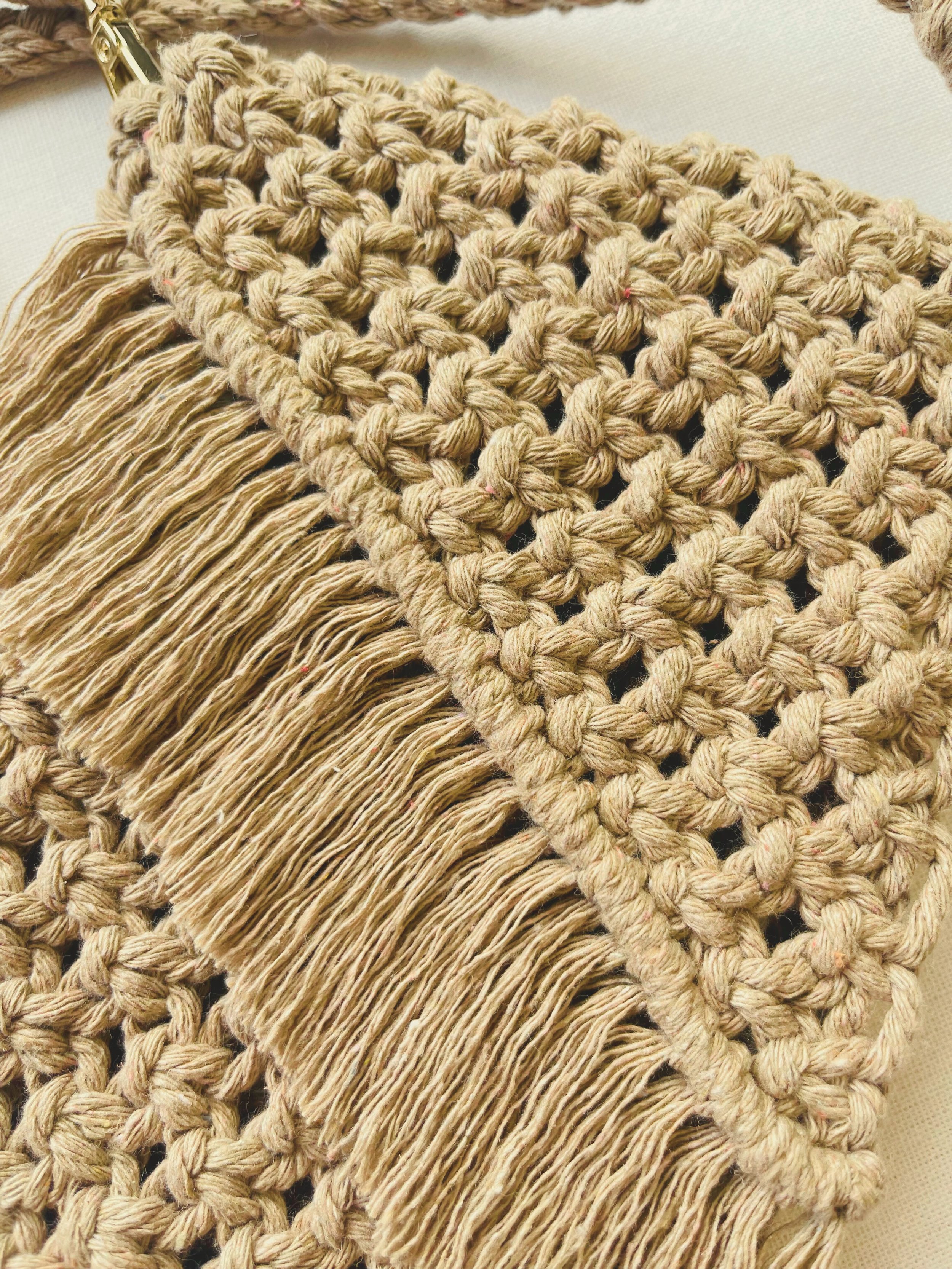 Light brown handmade macramé bag with diagonal fringe and detailed knot pattern, shown flat on an off-white fabric background.