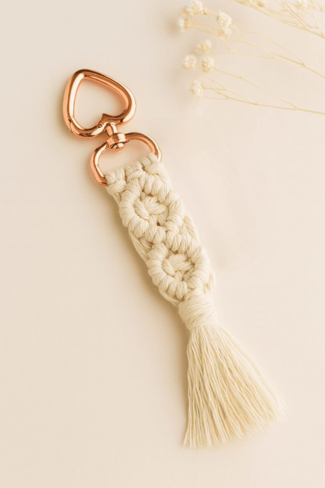 Off-white handmade macramé keychain with rose gold heart clasp, laid on a soft neutral background with dried baby's breath