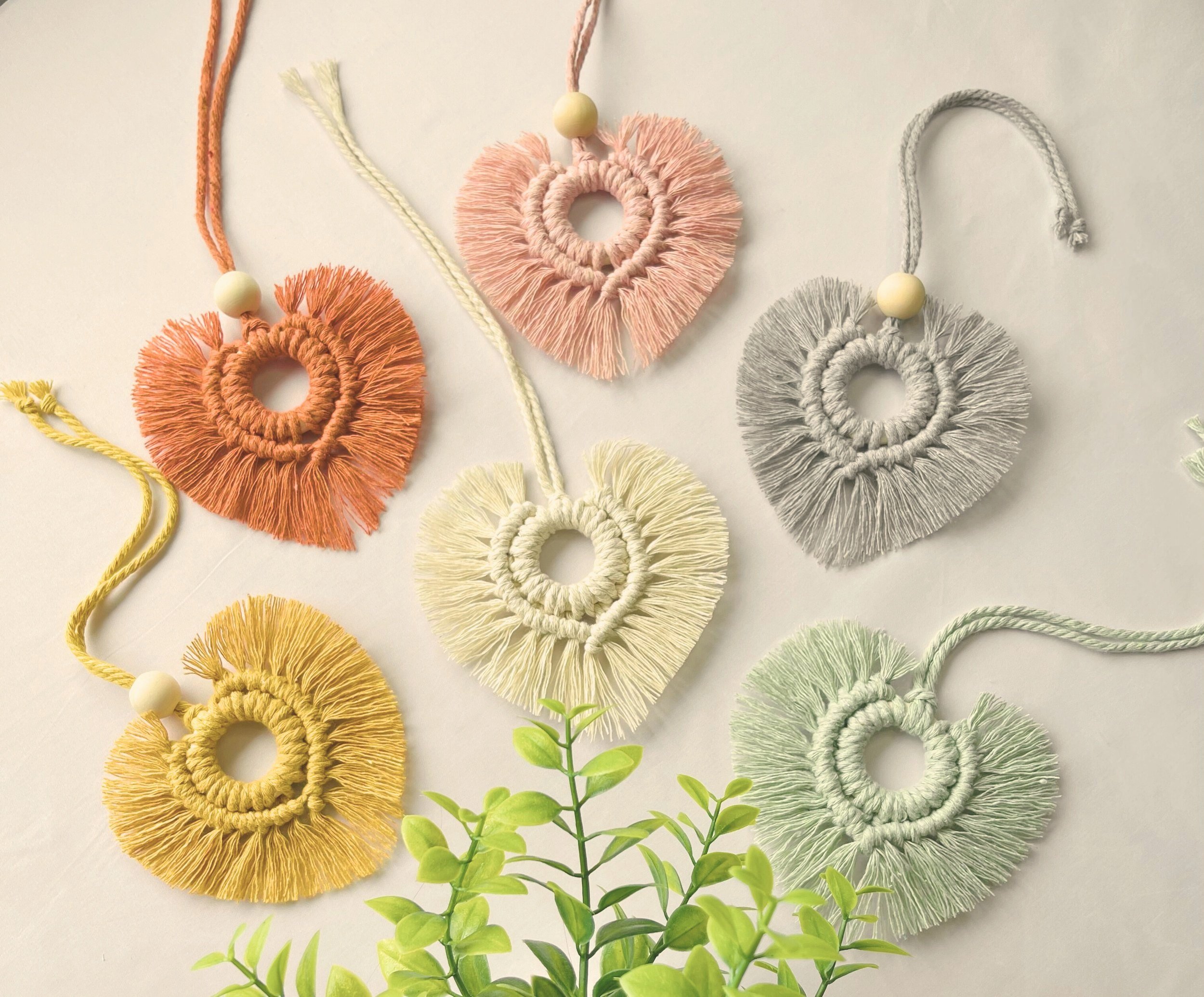 Six pastel macramé heart-shaped car diffusers with wood beads, styled on a soft off-white background with greenery – handmade by Samra Knots.