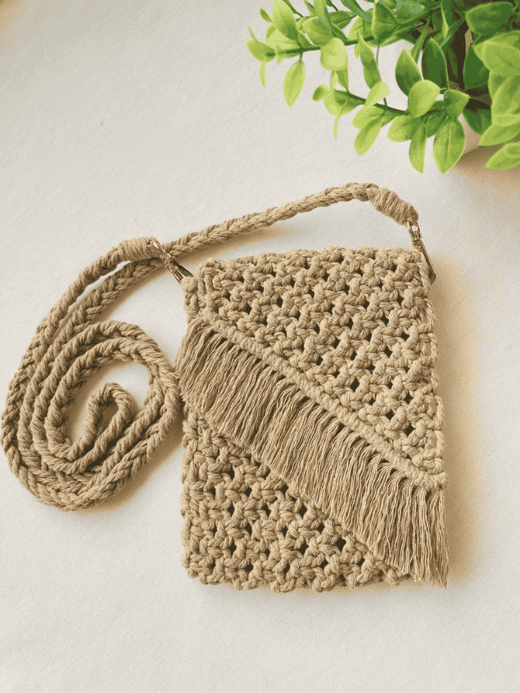 Full flat-lay of a light brown macramé crossbody bag with braided strap and fringe, displayed with green plant decor on neutral background.