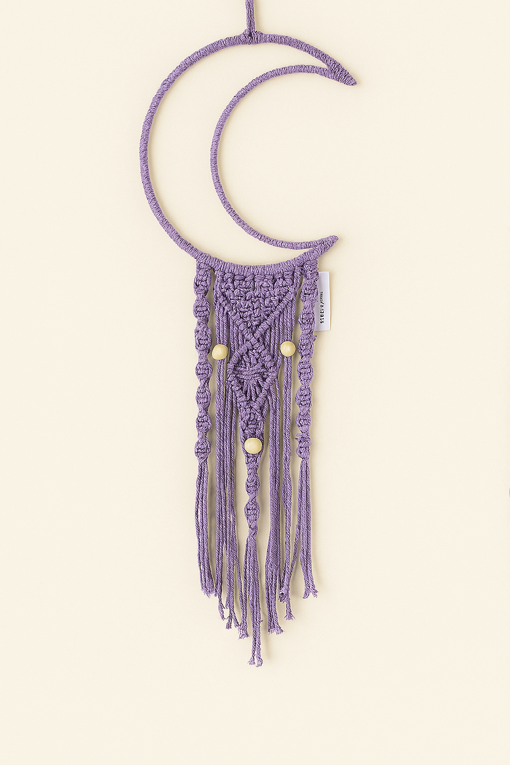 Handmade macramé crescent moon wall hanging in muted purple with natural wooden beads, displayed on a soft off-white background. Boho-style nursery or bedroom decor.