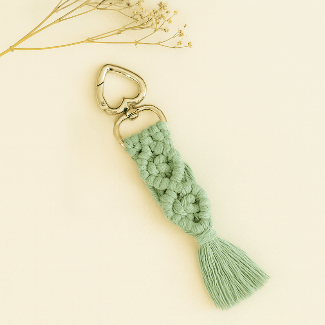 Soft sage green macramé keychain with silver heart clasp, displayed on a neutral background with a sprig of dried flowers