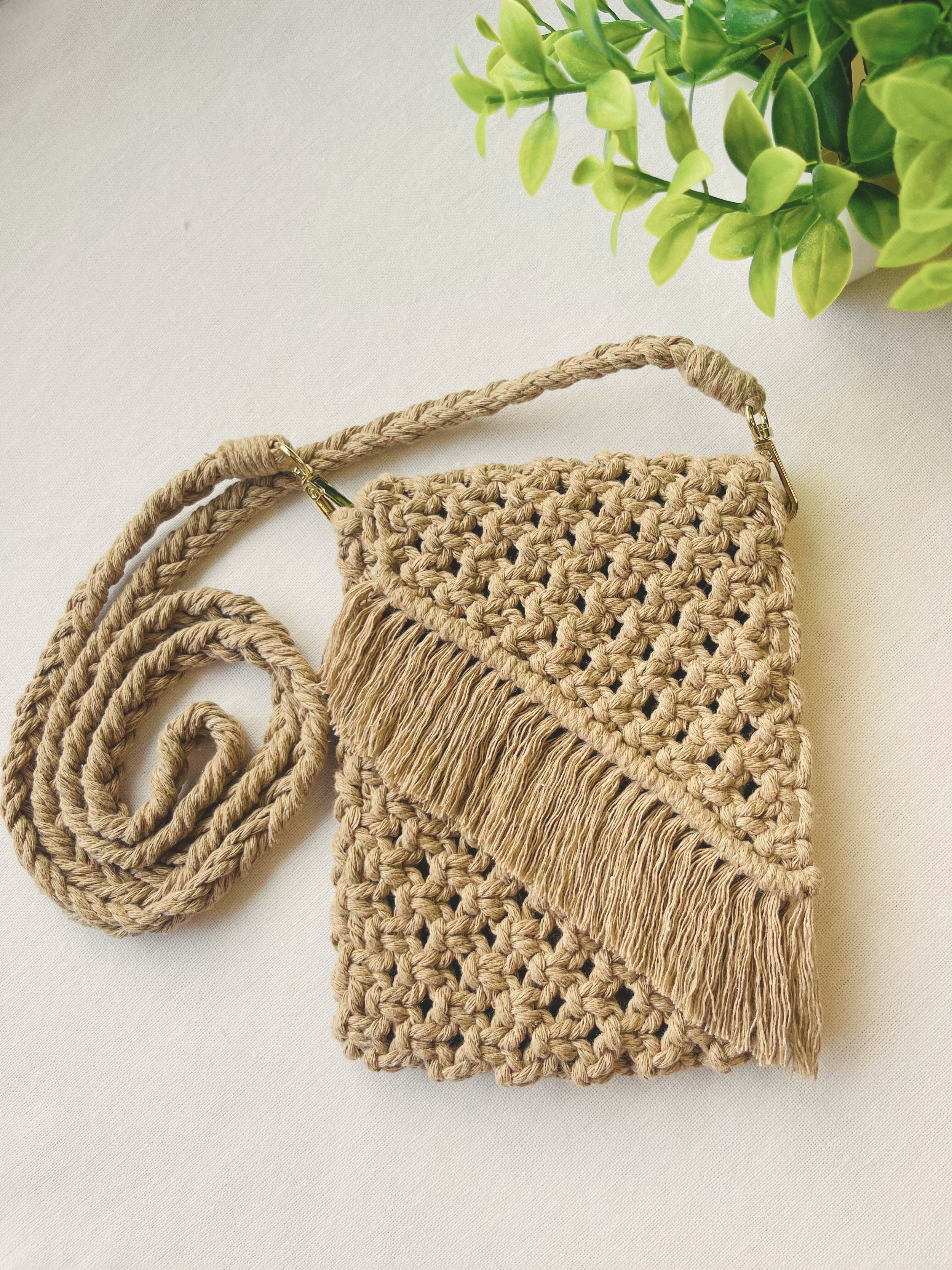 Full flat-lay of a light brown macramé crossbody bag with braided strap and fringe, displayed with green plant decor on neutral background.