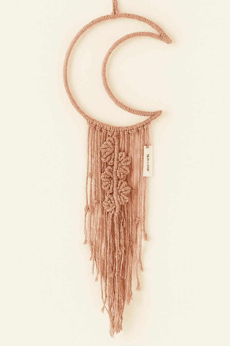 Boho macramé wall hanging shaped like a crescent moon in dusty pink, featuring floral-inspired knots and long fringe. Neutral backdrop, perfect for cozy home decor.