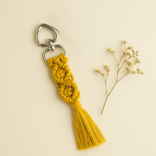Handmade macramé diamond keychain in mustard yellow with silver heart clasp, styled on an off-white background with dried flowers